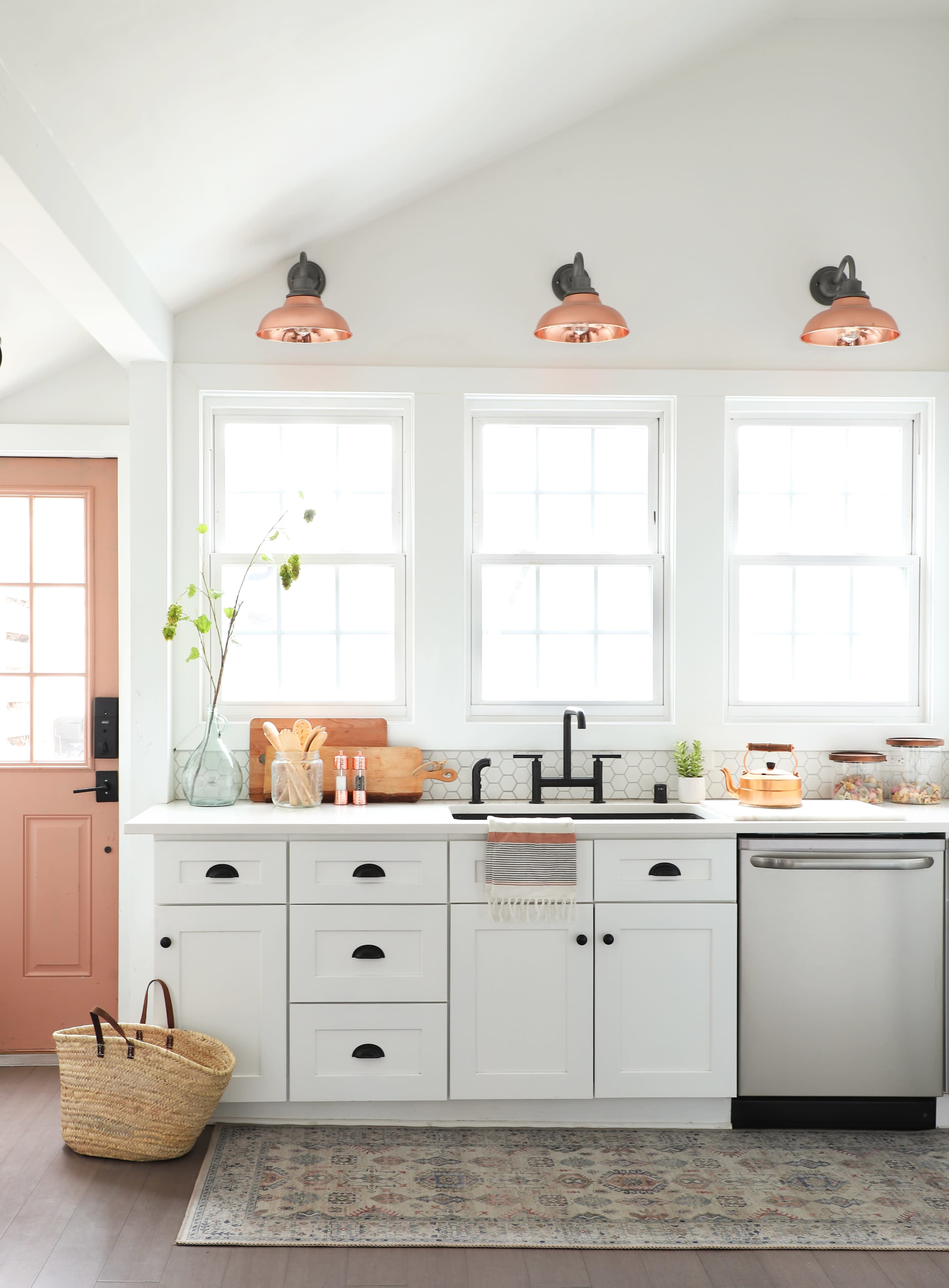 The kitchen features white cabinetry, a stainless steel dishwasher, a black faucet, and large windows with copper light fixtures above.