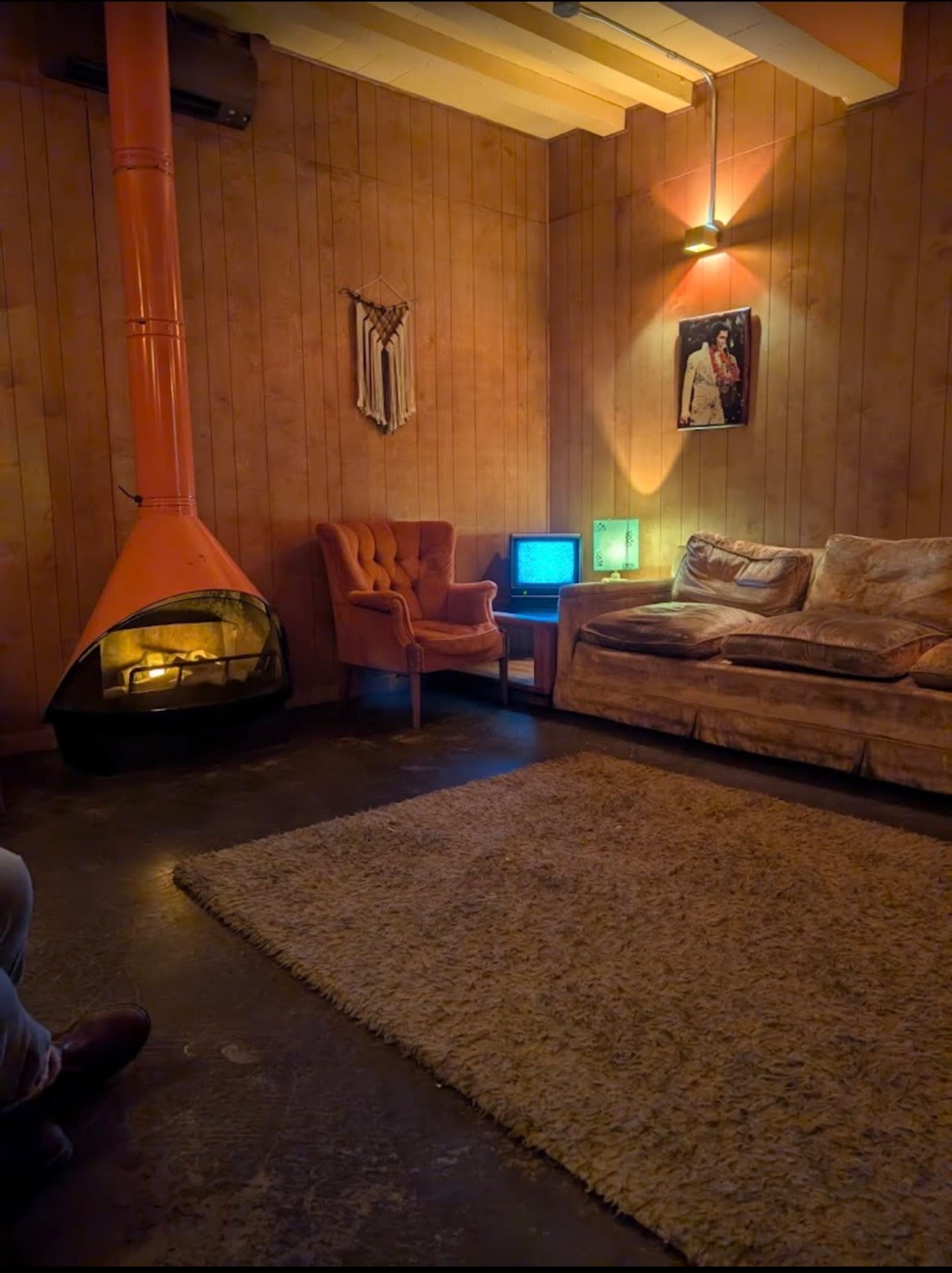 The image shows a cozy living room with a retro orange fireplace, a plush couch, an armchair, and a vintage television, all set against wooden paneling.