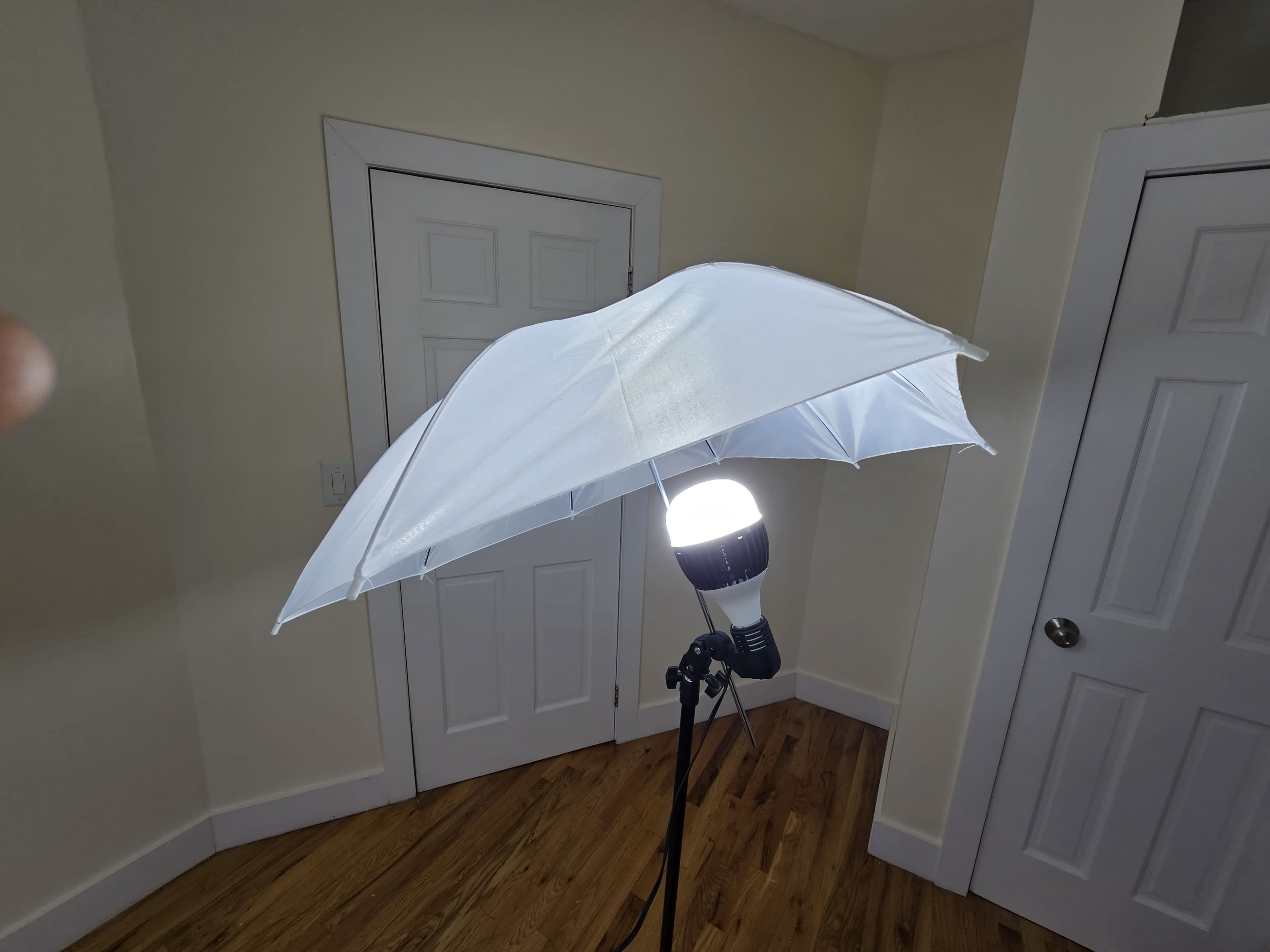 A white umbrella is mounted on a stand, above a bright light bulb, positioned in a room with wooden flooring and a beige wall.