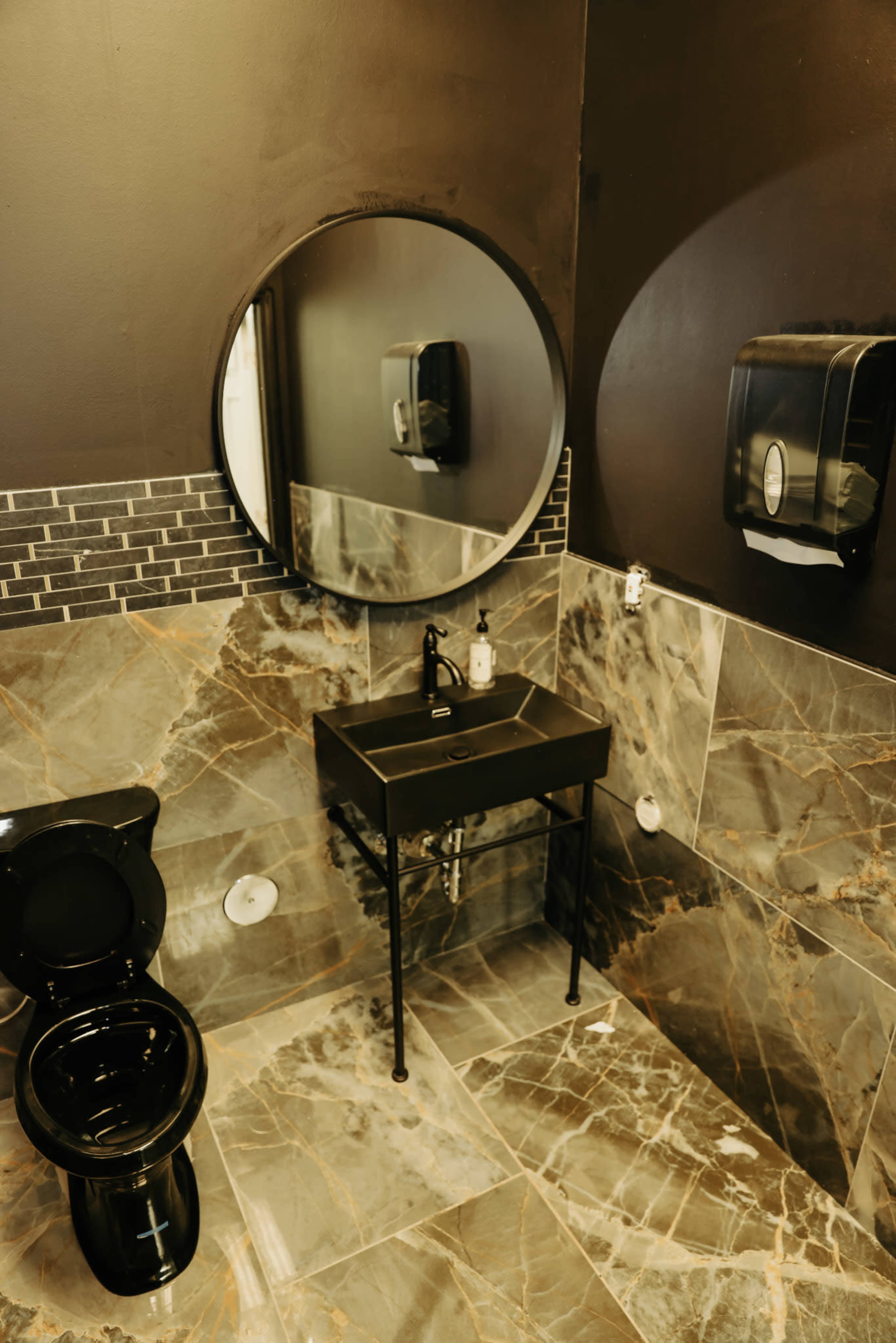 The image shows a modern bathroom featuring a black toilet, a round mirror, a sleek sink with a faucet, and marble-patterned tiles on the walls and floor.