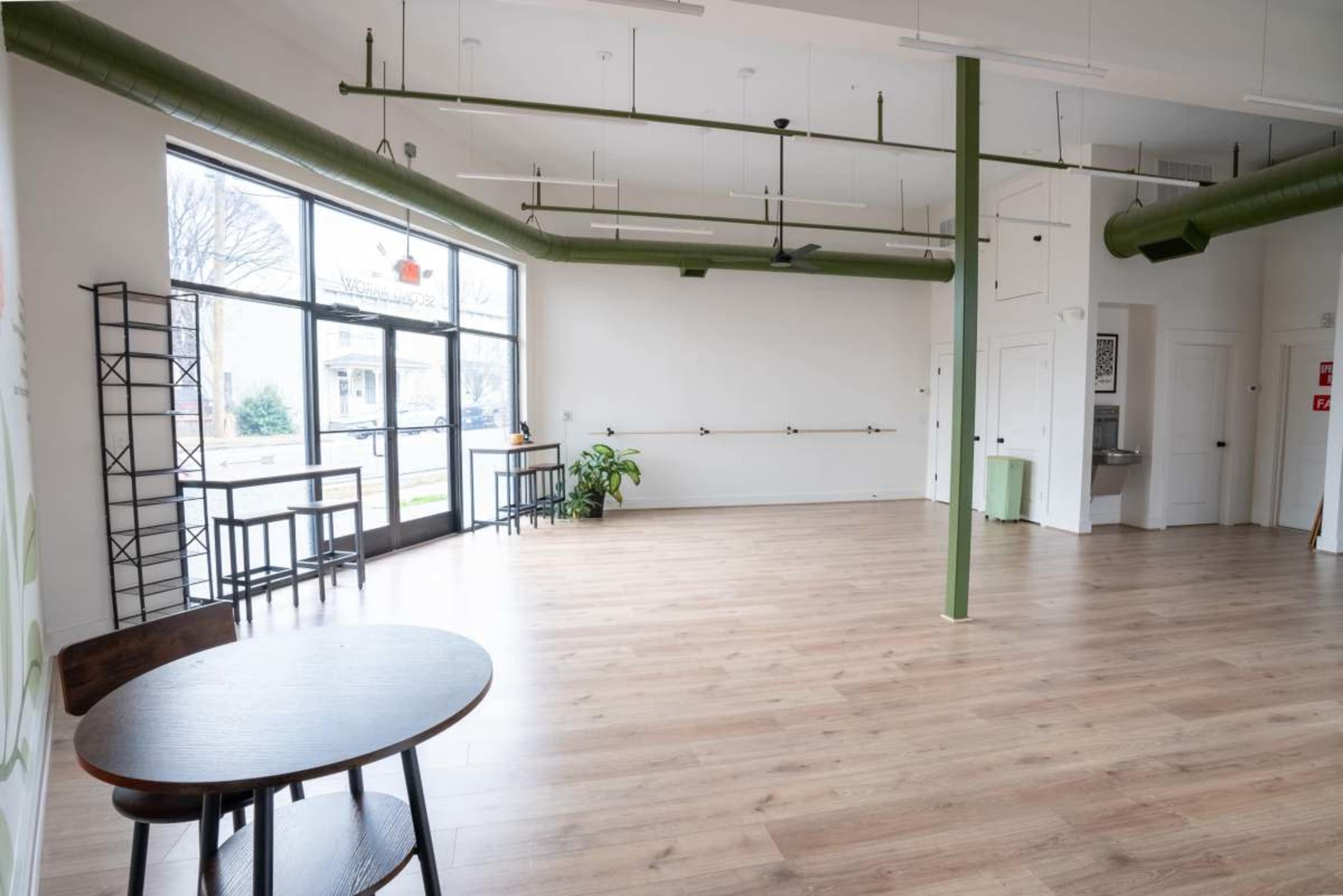 The image shows a spacious, well-lit room with wooden flooring, featuring a few tables, green accent pipes, and large windows that allow natural light to enter.