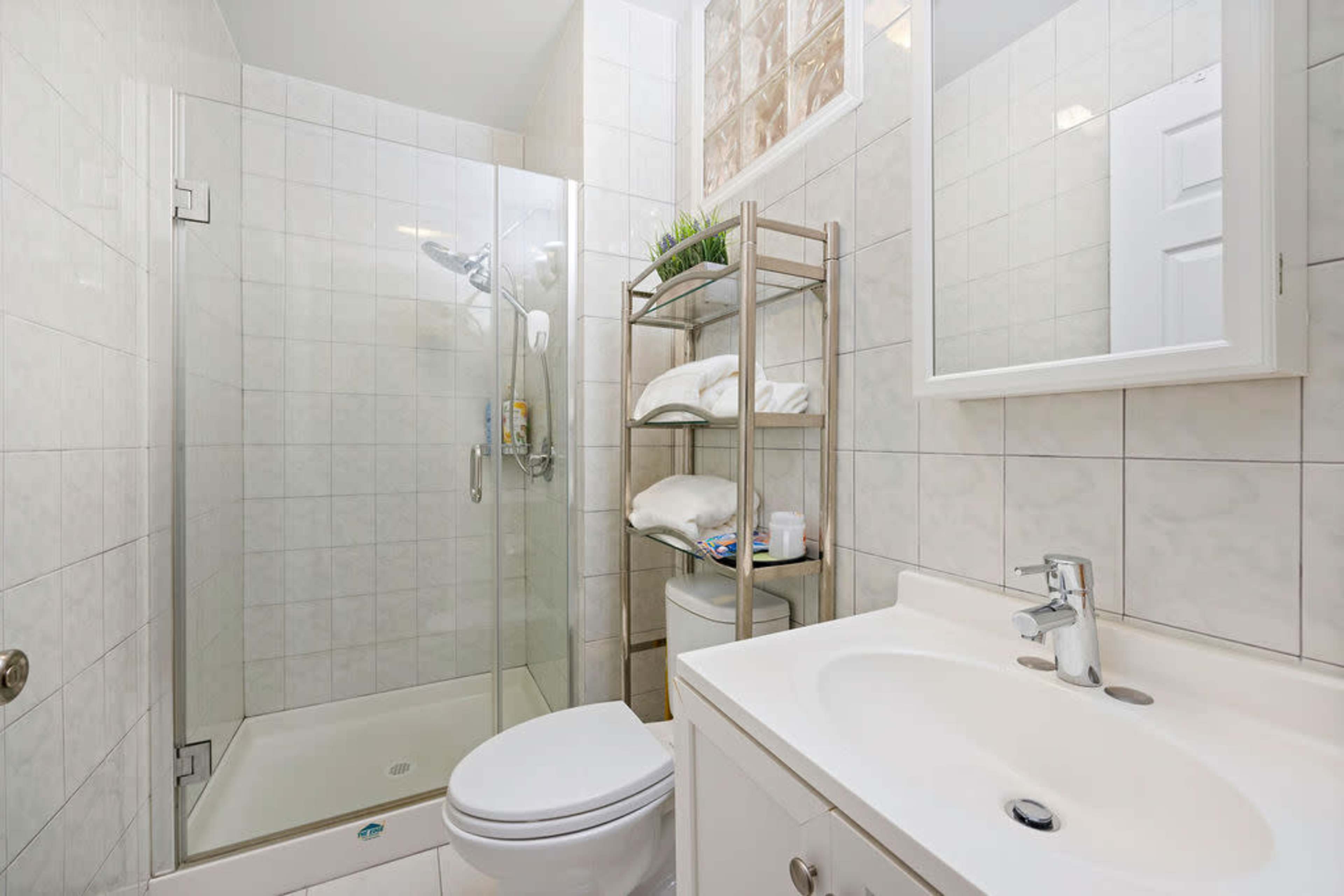 A modern bathroom features a glass shower, a toilet, and a sink with a mirror, along with a metal shelf holding towels and toiletries.