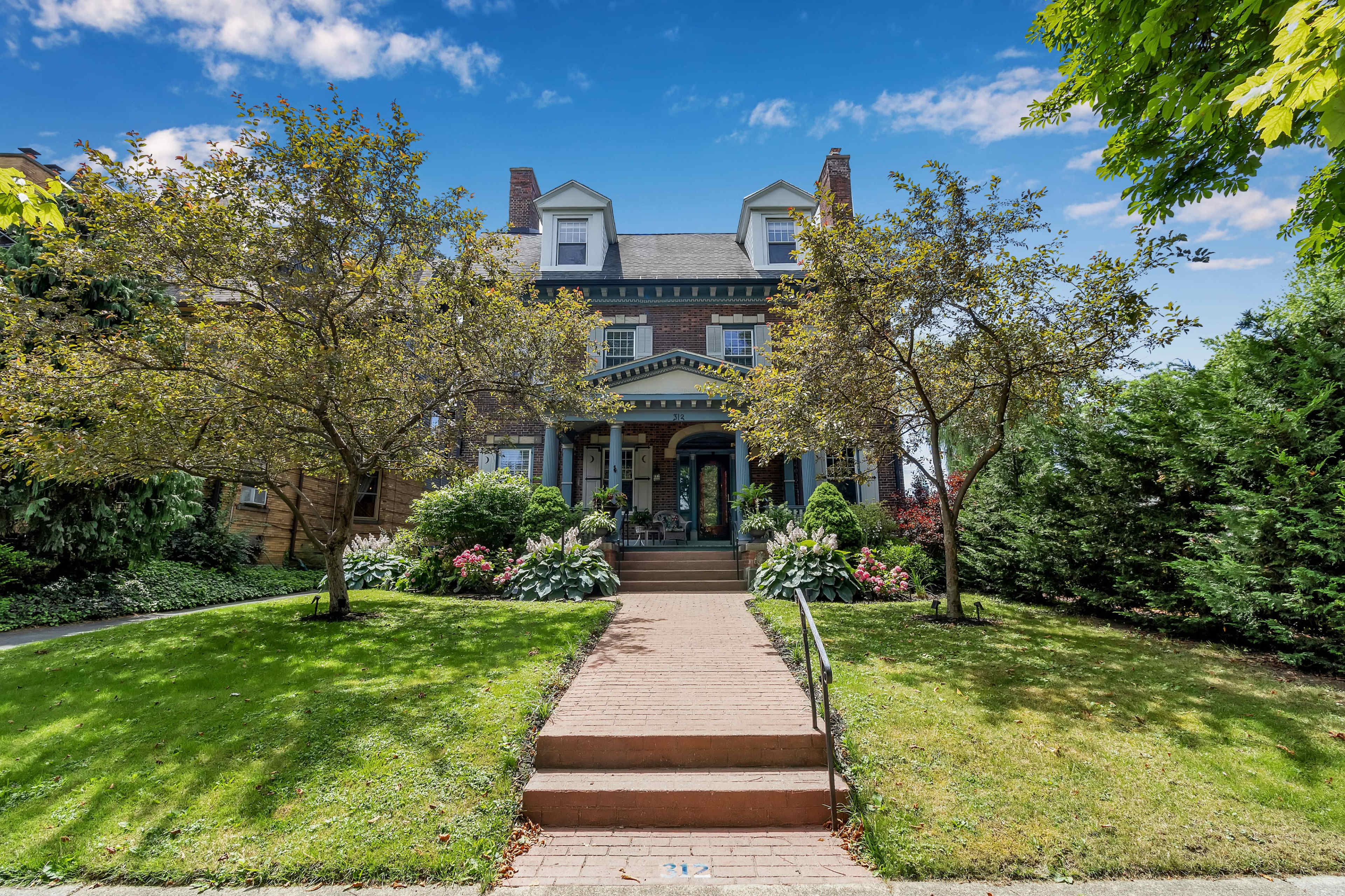 A large historic house with a front porch is framed by well-manicured lawns and trees, leading up to a set of steps.