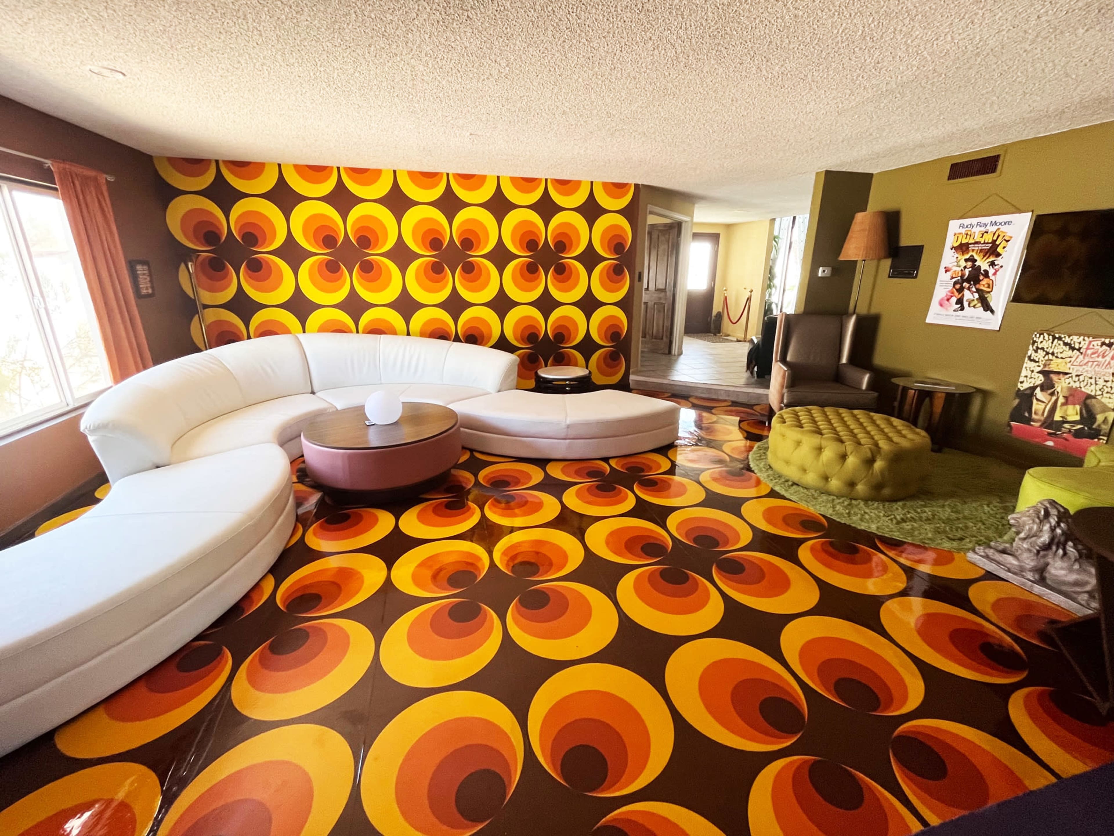A living room with a curved white sofa, a circular coffee table, and a boldly patterned yellow and brown floor, accompanied by colorful wallpaper featuring concentric circles.