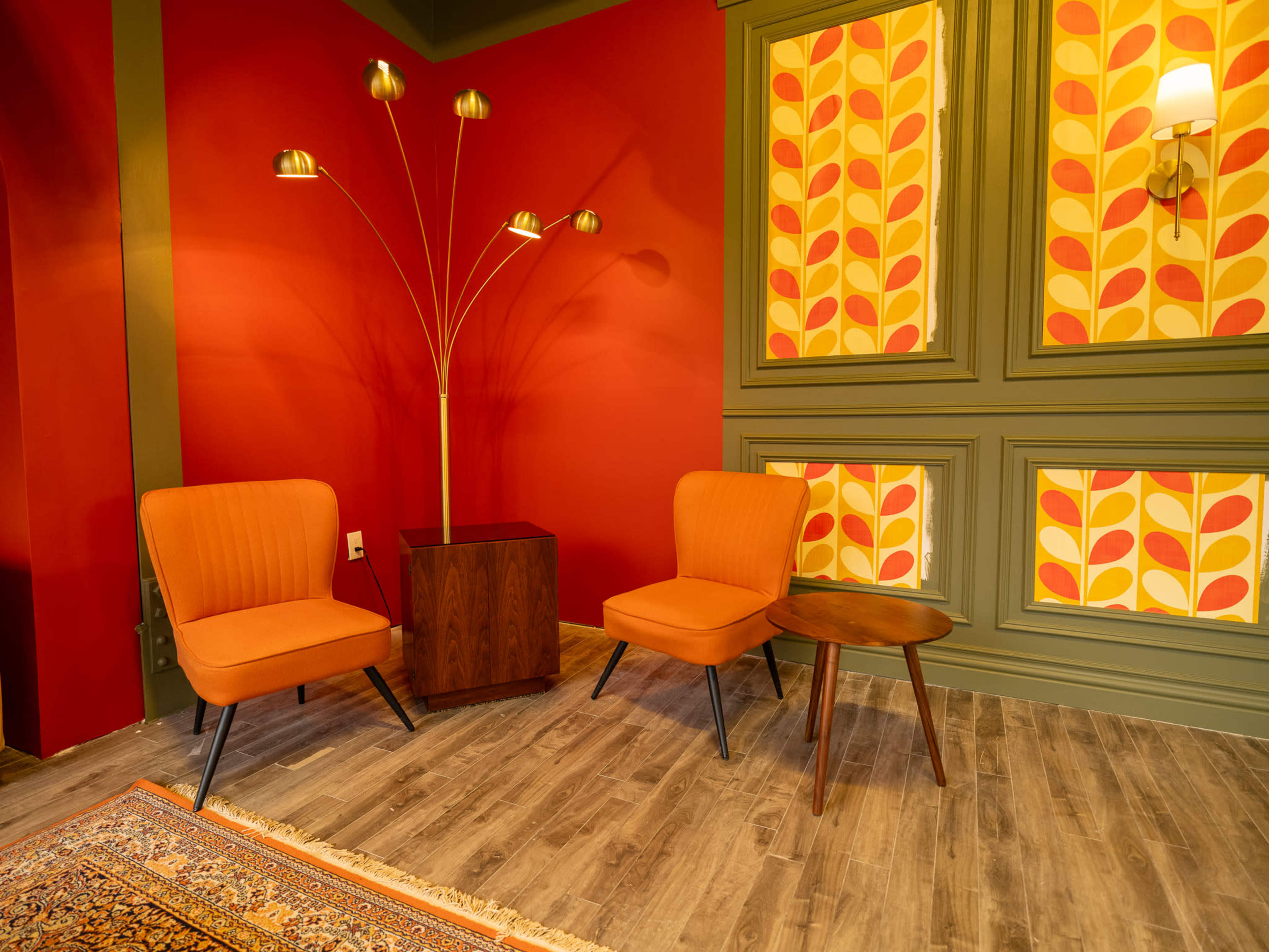 The scene features two orange armchairs and a small wooden table against a red wall decorated with patterned wallpaper.