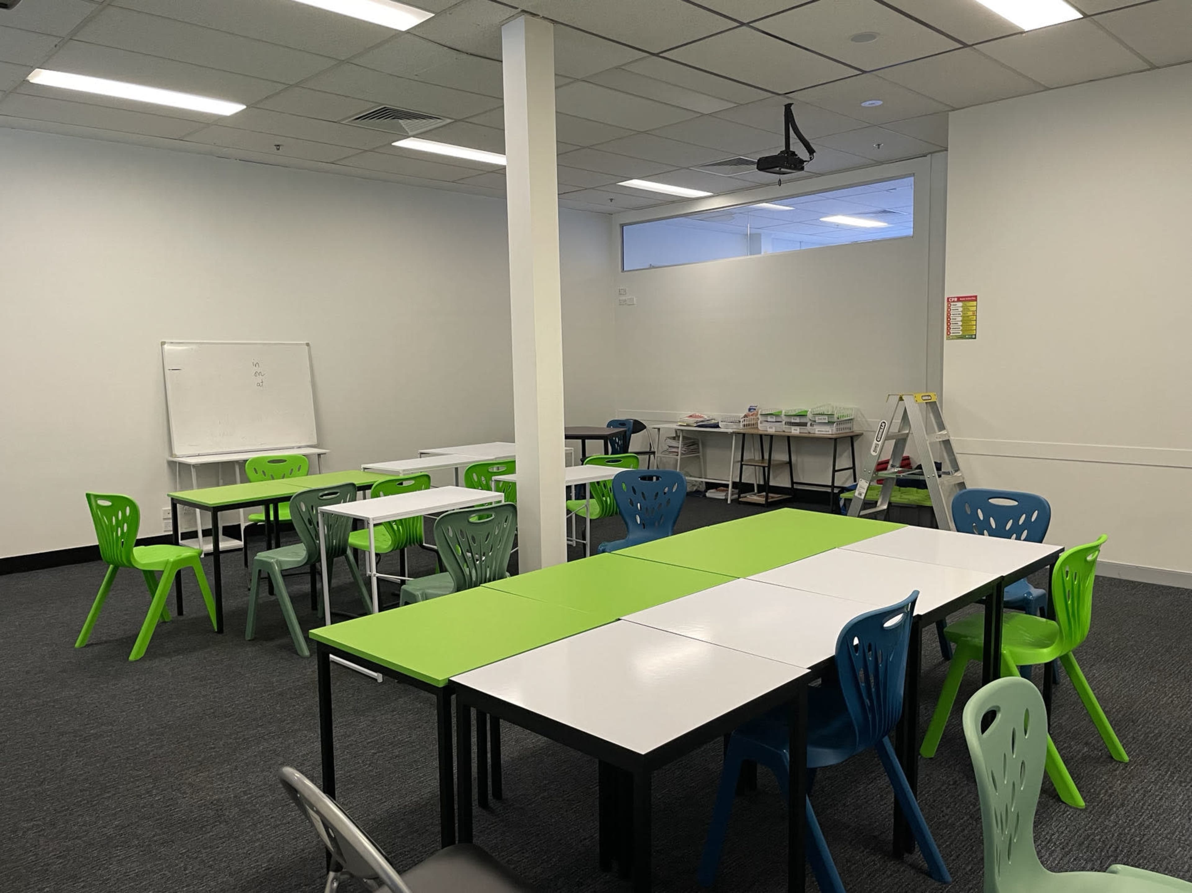 A classroom setting features multiple tables with green and white surfaces, accompanied by colorful chairs and a whiteboard, all arranged in a spacious area.