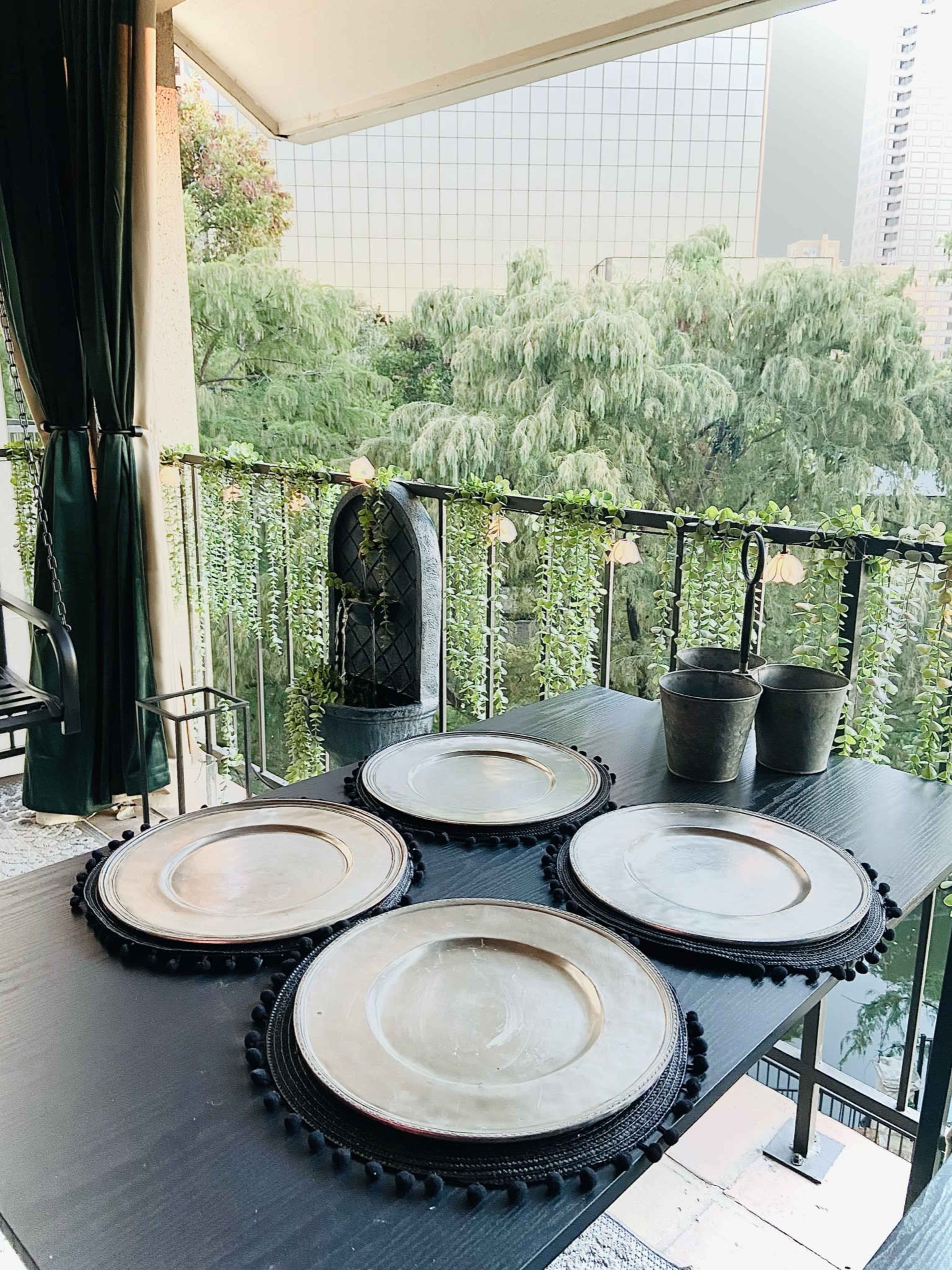 A set of round, metallic plates is arranged on a dark table with greenery and buildings visible in the background.