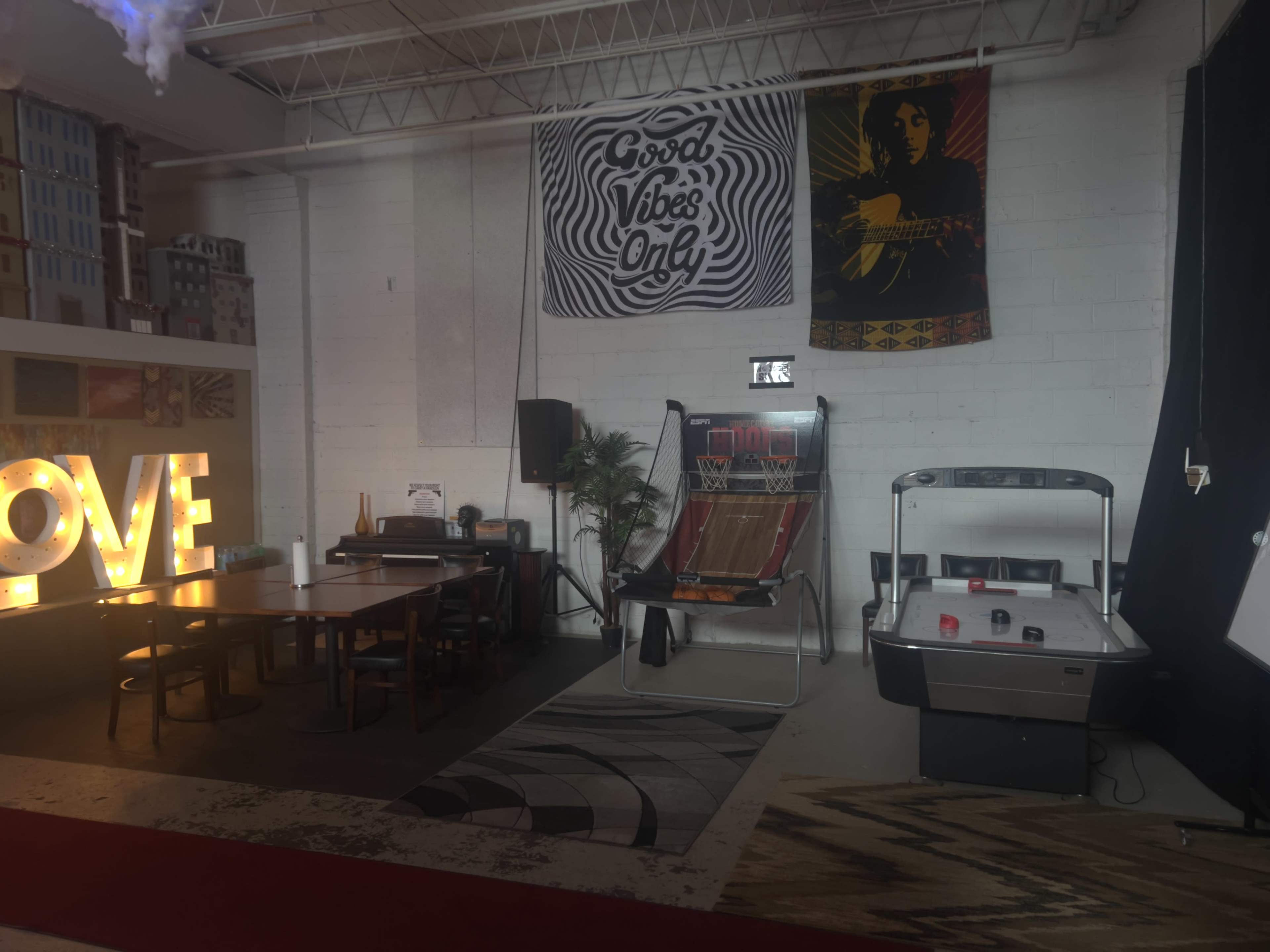 A recreational space featuring a large illuminated "LOVE" sign, a wooden dining table, an air hockey table, and wall decorations including a tapestry with the phrase "Good Vibes Only."