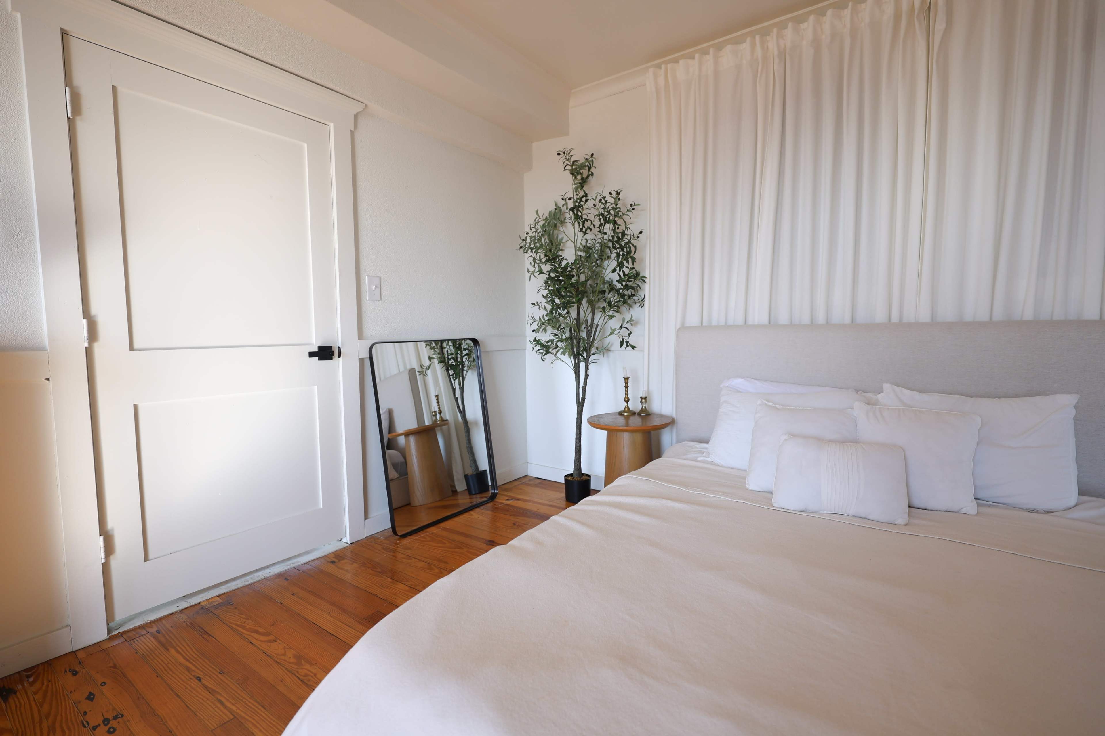 The image shows a minimalistic bedroom featuring a bed with white bedding, a large mirror, a potted plant, and a closed white door.