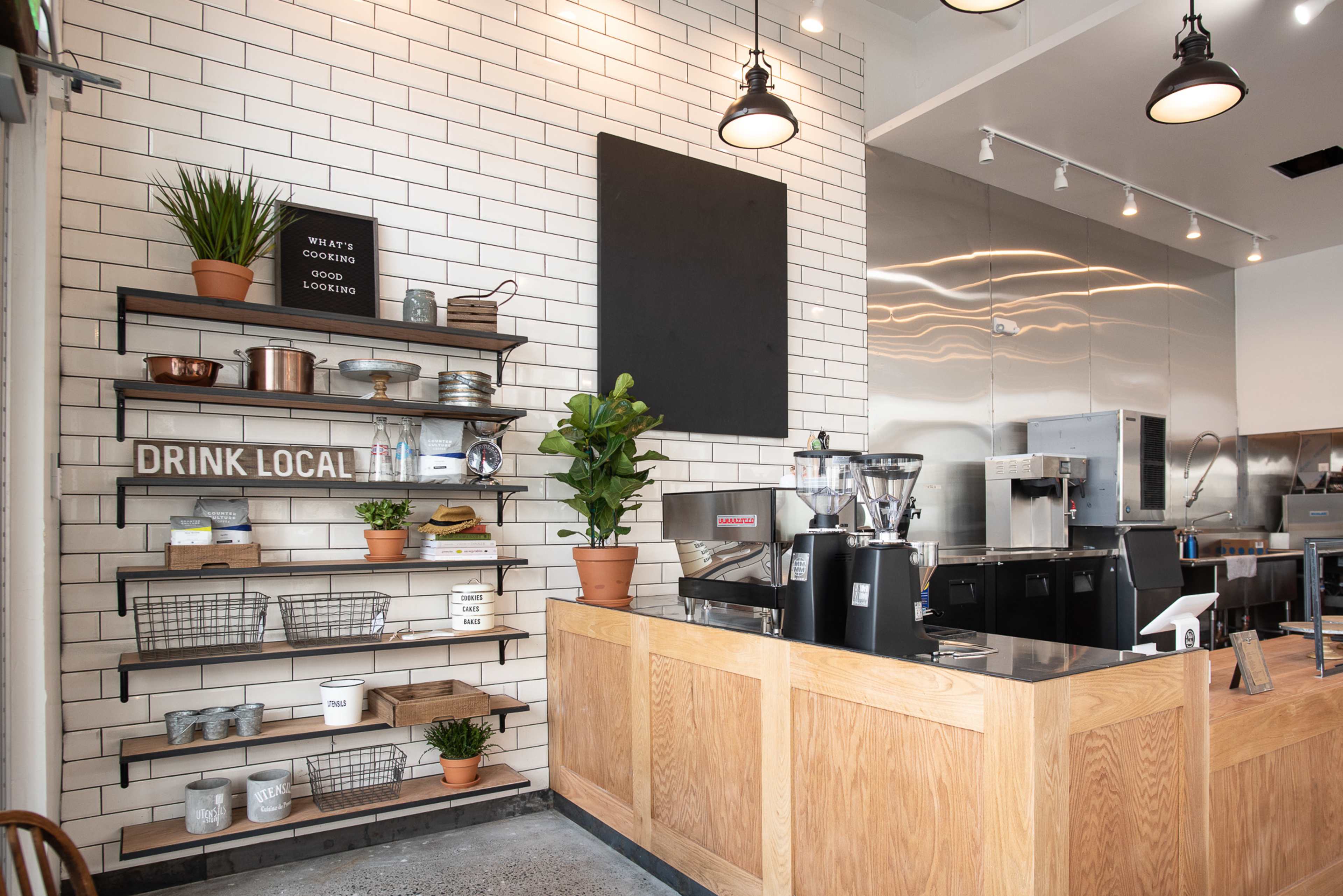 A coffee shop interior with a wooden counter, metal shelving displaying various kitchen items, and a clean, tiled wall.