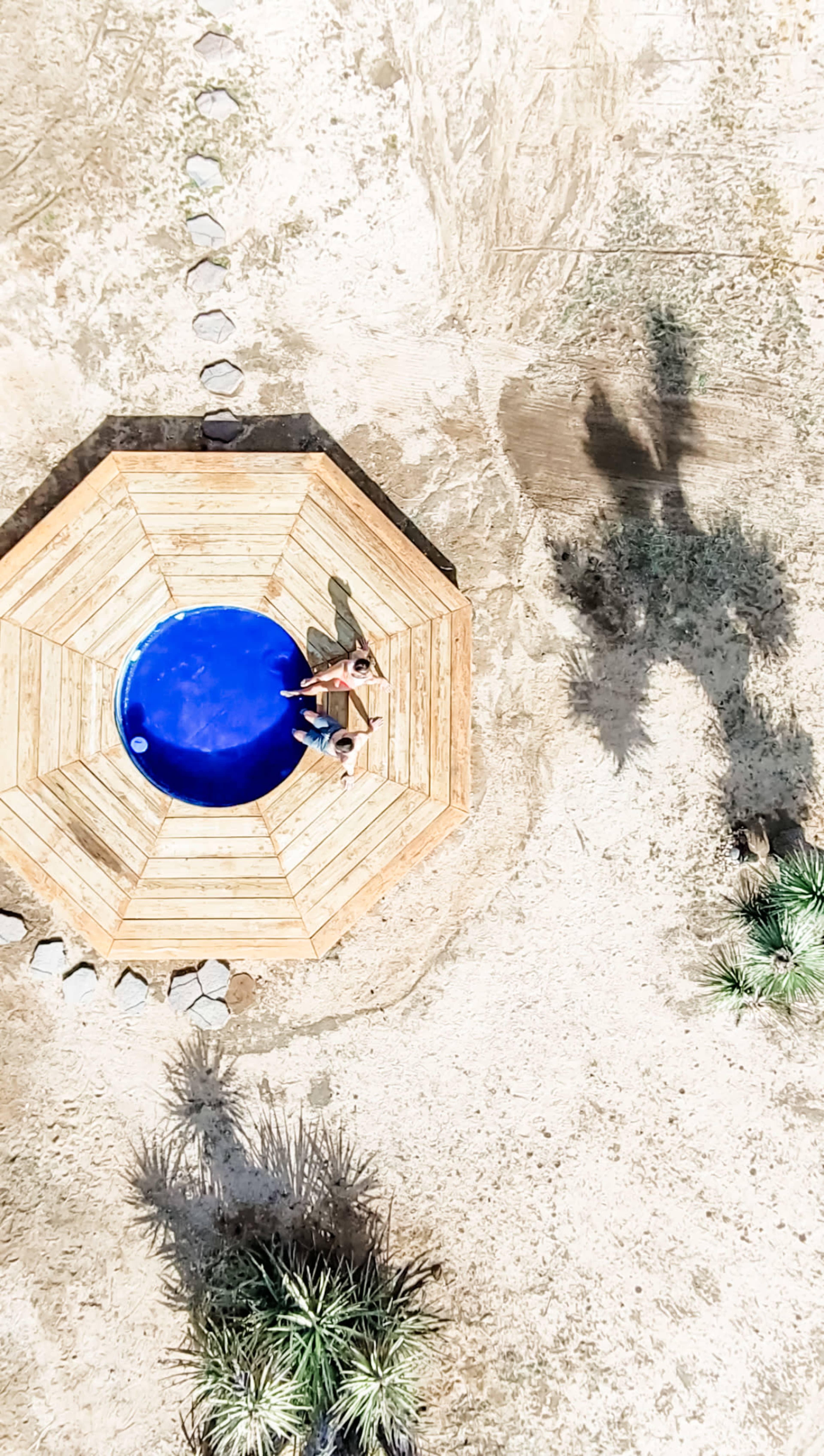 Two people sit on a wooden deck around a circular blue pool surrounded by dry ground and sparse vegetation.