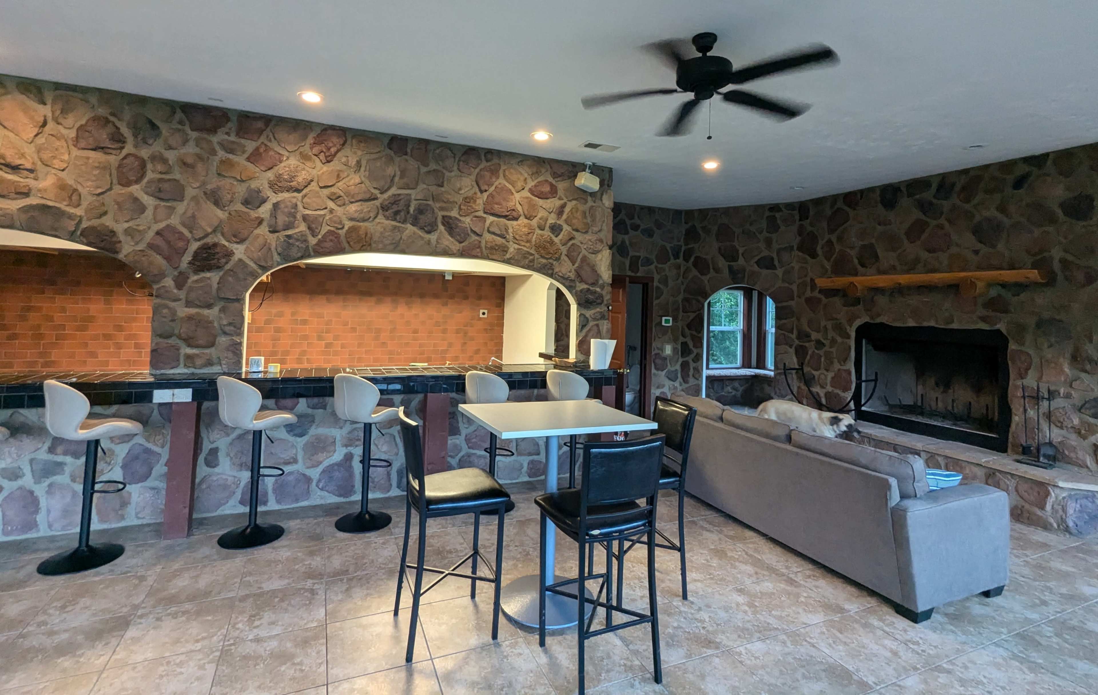 A spacious indoor area featuring a stone wall, a bar with stools, a table, and a couch with a fireplace in the background.