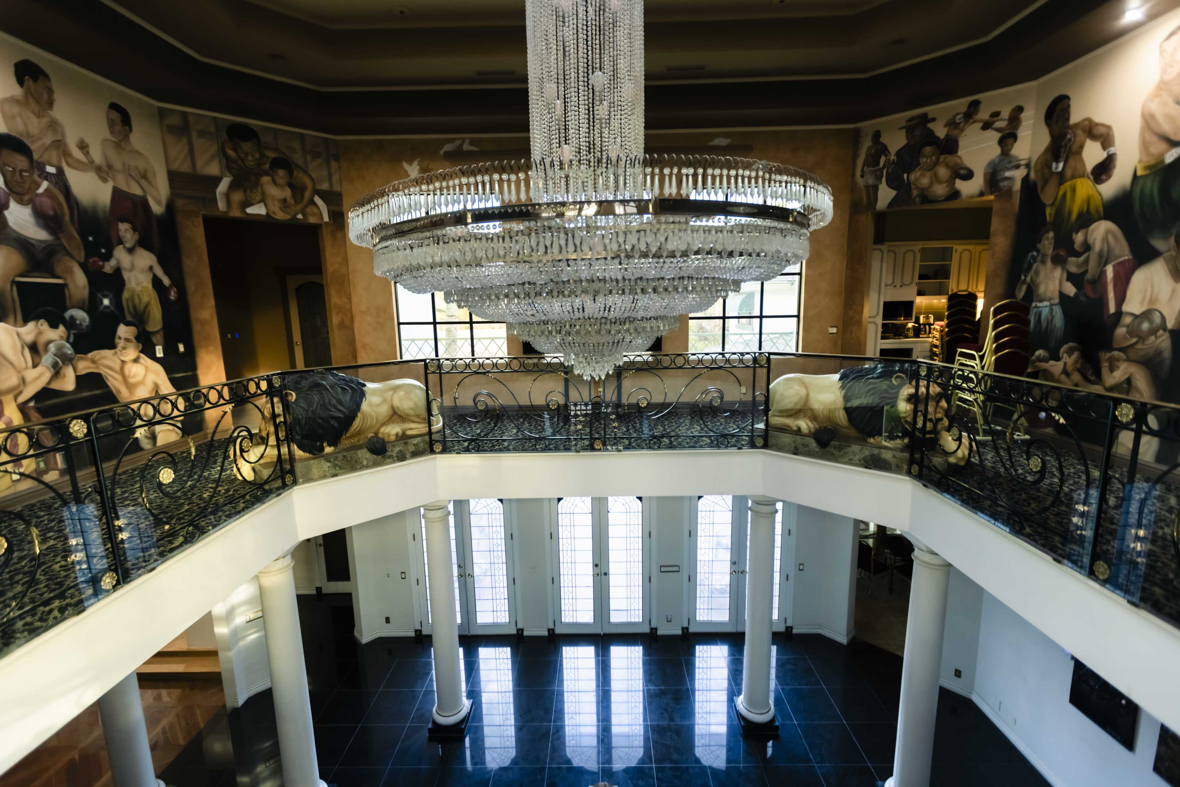 The image shows a luxurious interior of a mansion featuring a grand chandelier, intricate railings, and large murals of boxers on the walls.