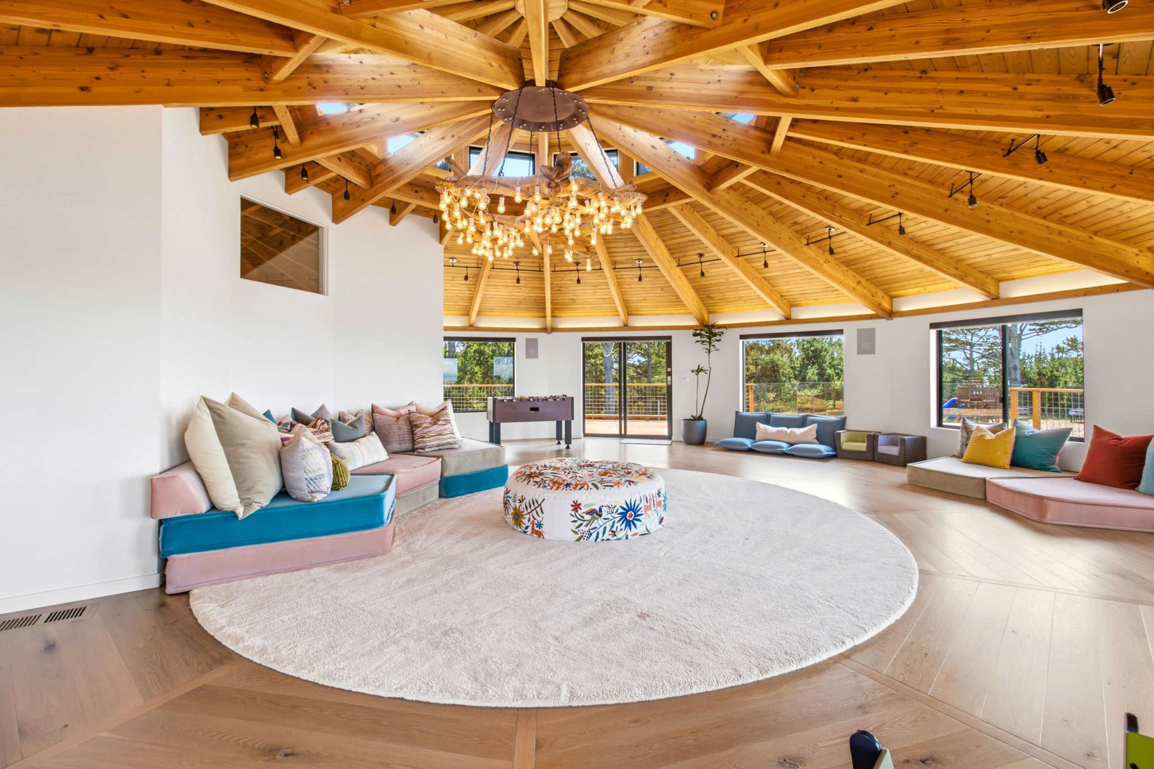 The room features a circular layout with a wooden ceiling, large windows, and a central round rug surrounded by cushioned seating areas.