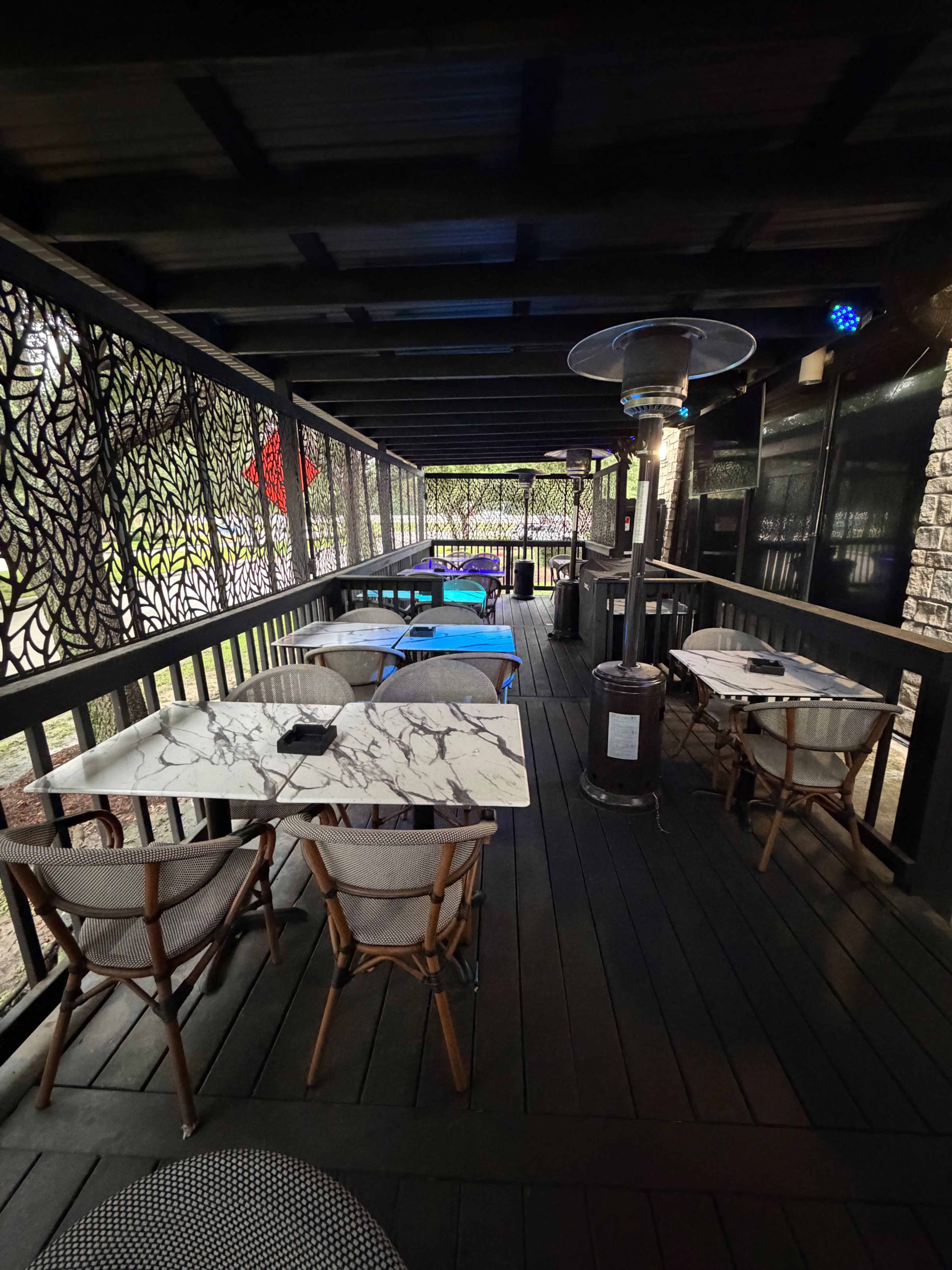 The image shows a covered outdoor dining area with tables and chairs arranged along a wooden deck, partially enclosed by decorative screens.