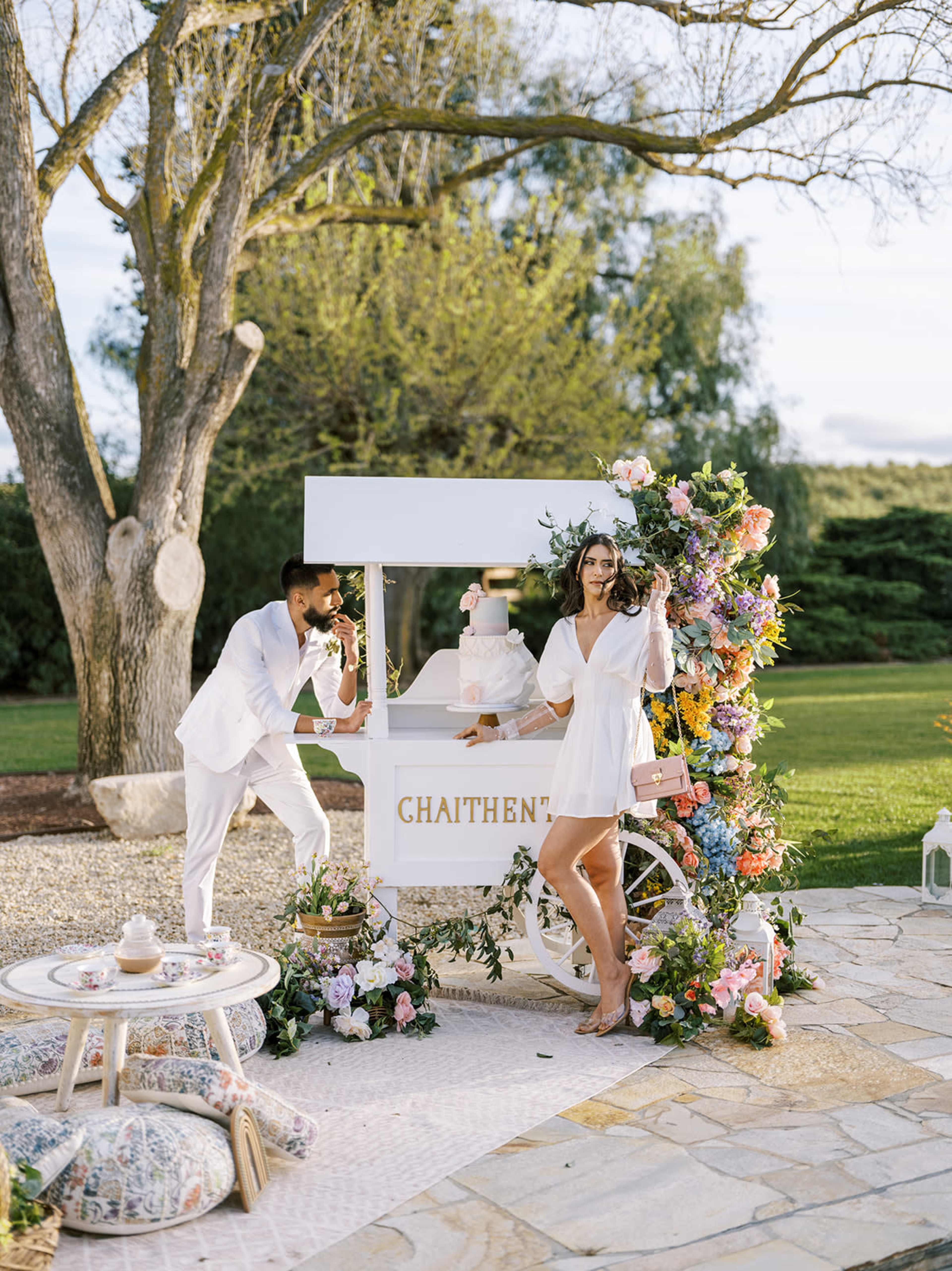 A couple poses beside a decorated ice cream cart surrounded by flowers in an outdoor setting.