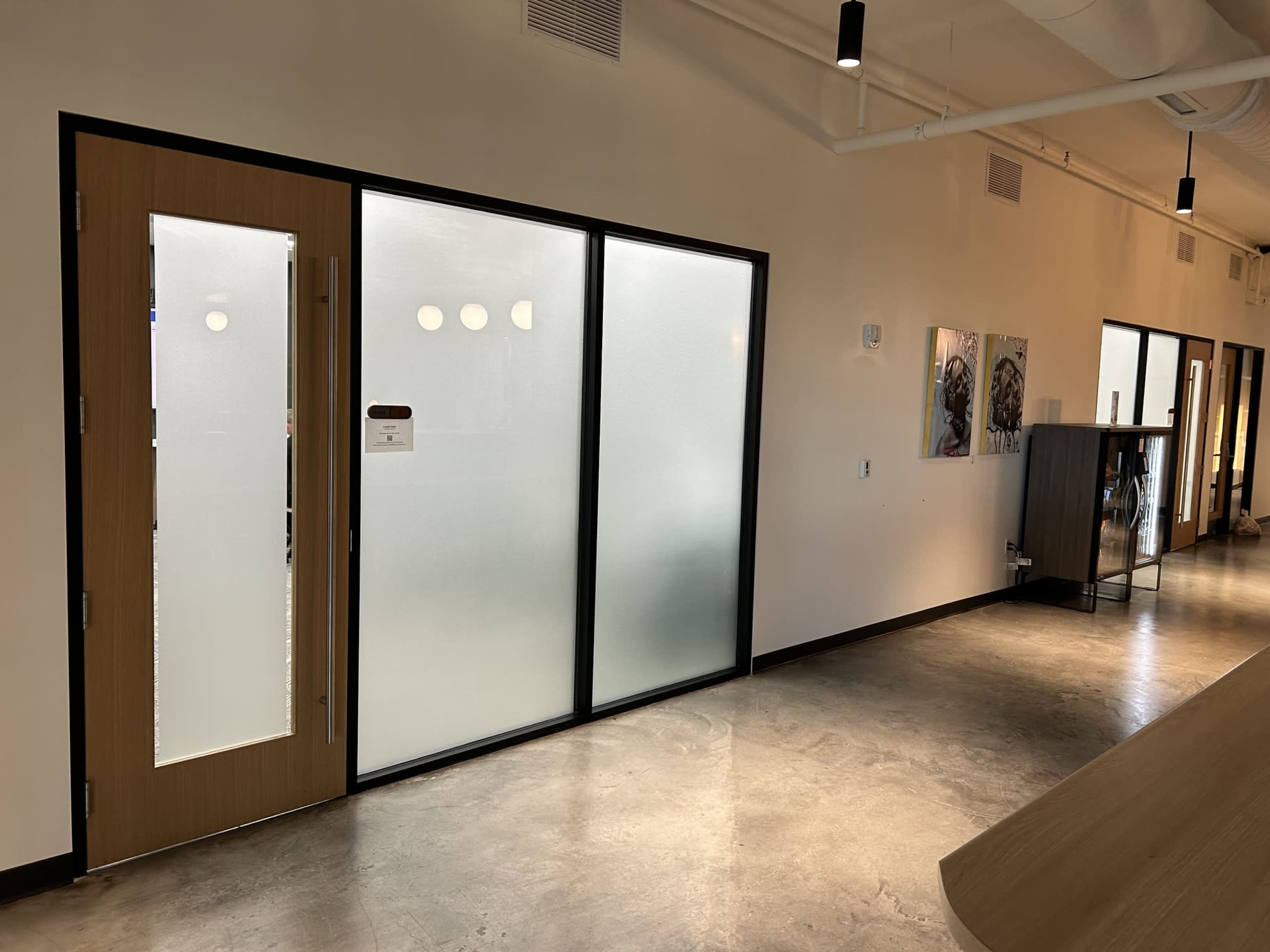 The image shows a modern office hallway with a glass door leading to a room and a sleek, minimalist design featuring concrete floors and contemporary lighting.