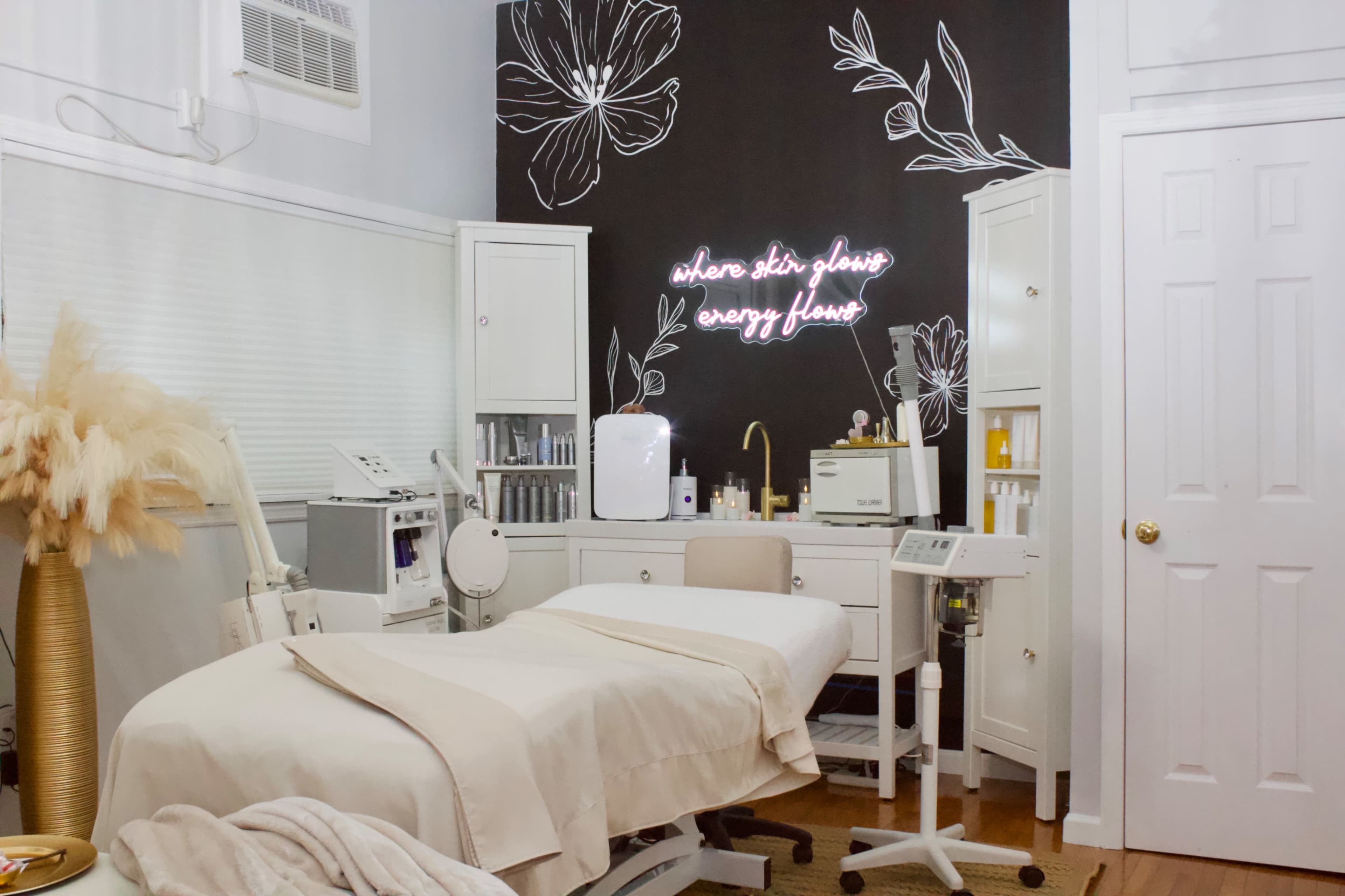 A skincare treatment room features a treatment bed, various skincare products, and a wall decorated with floral designs and a neon sign.
