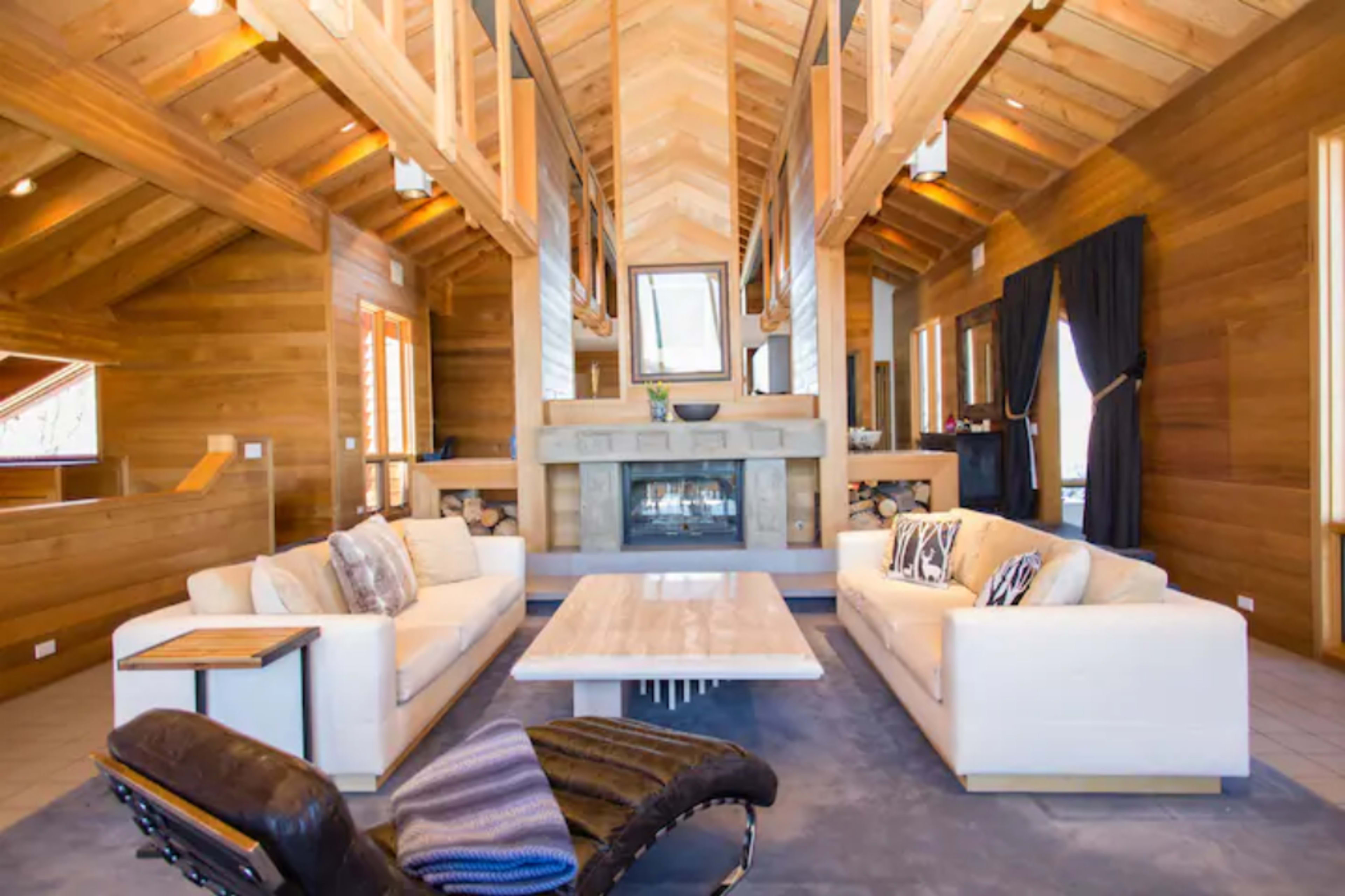 The living room features a high wooden ceiling, two white sofas facing a stone fireplace, and a light-colored coffee table.