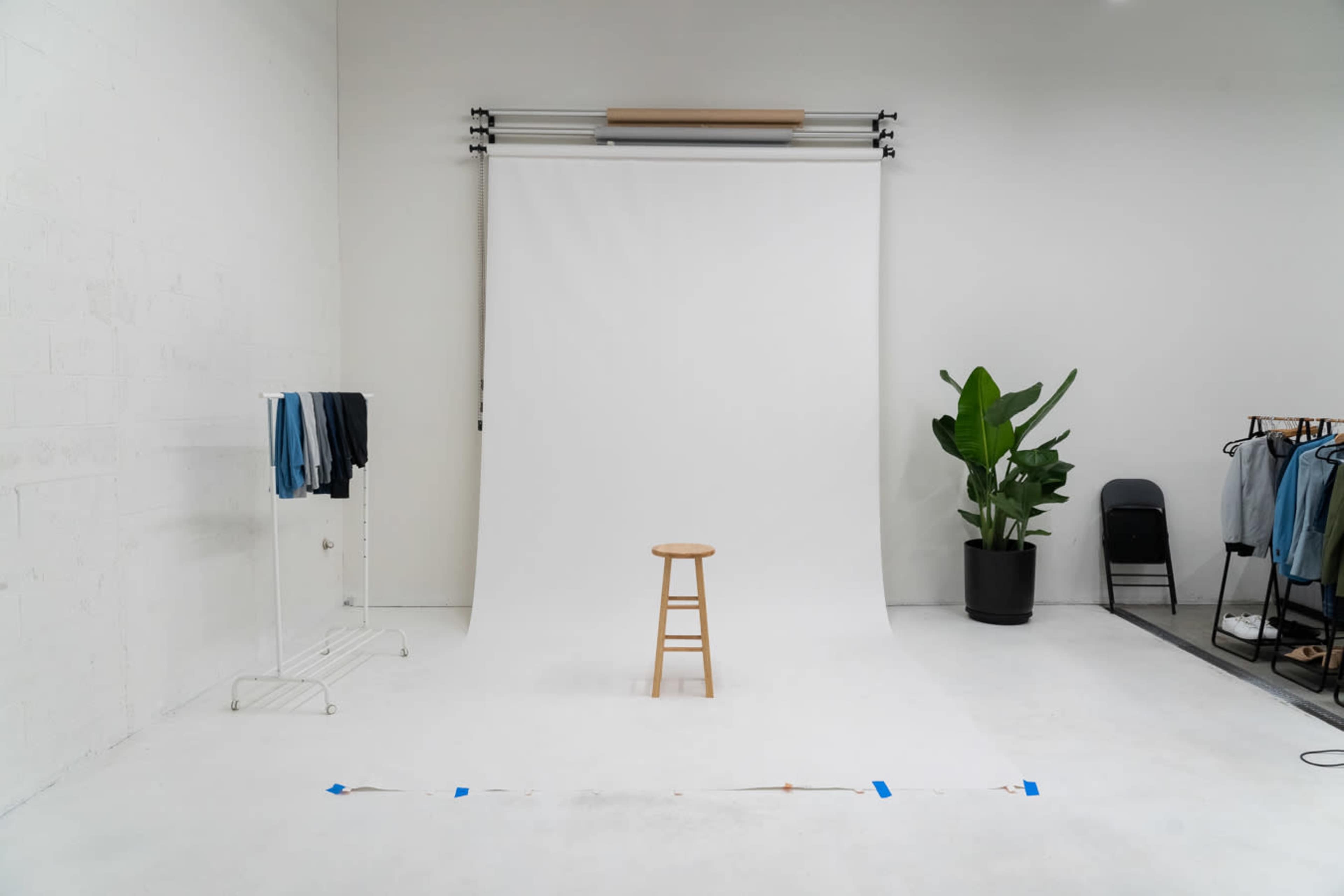 A simple studio setup features a white backdrop, a wooden stool in the center, and clothing displays on the sides.