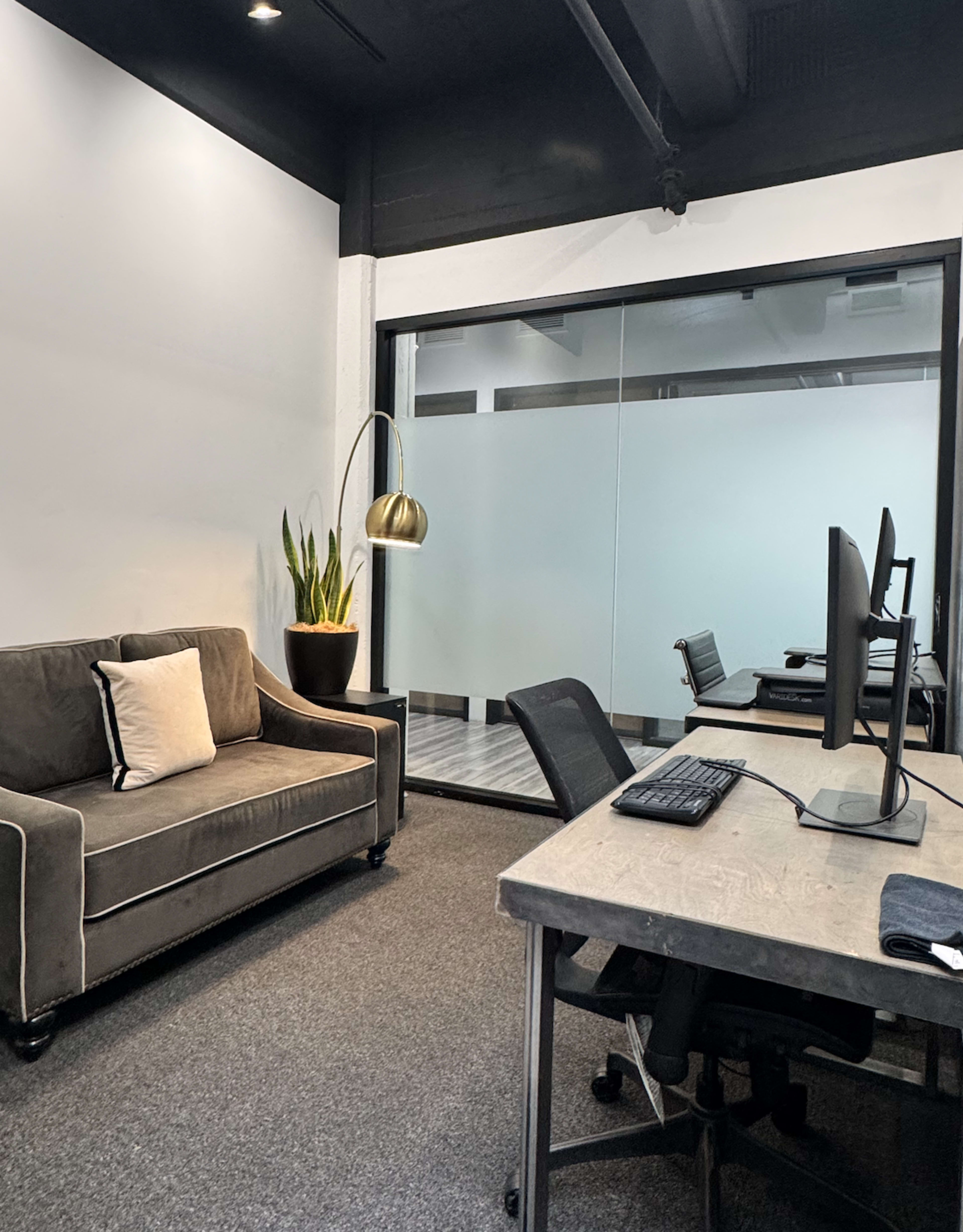 The image shows a modern office space featuring a brown sofa, a large plant in a pot, and two desks with computers facing a glass-walled meeting room.