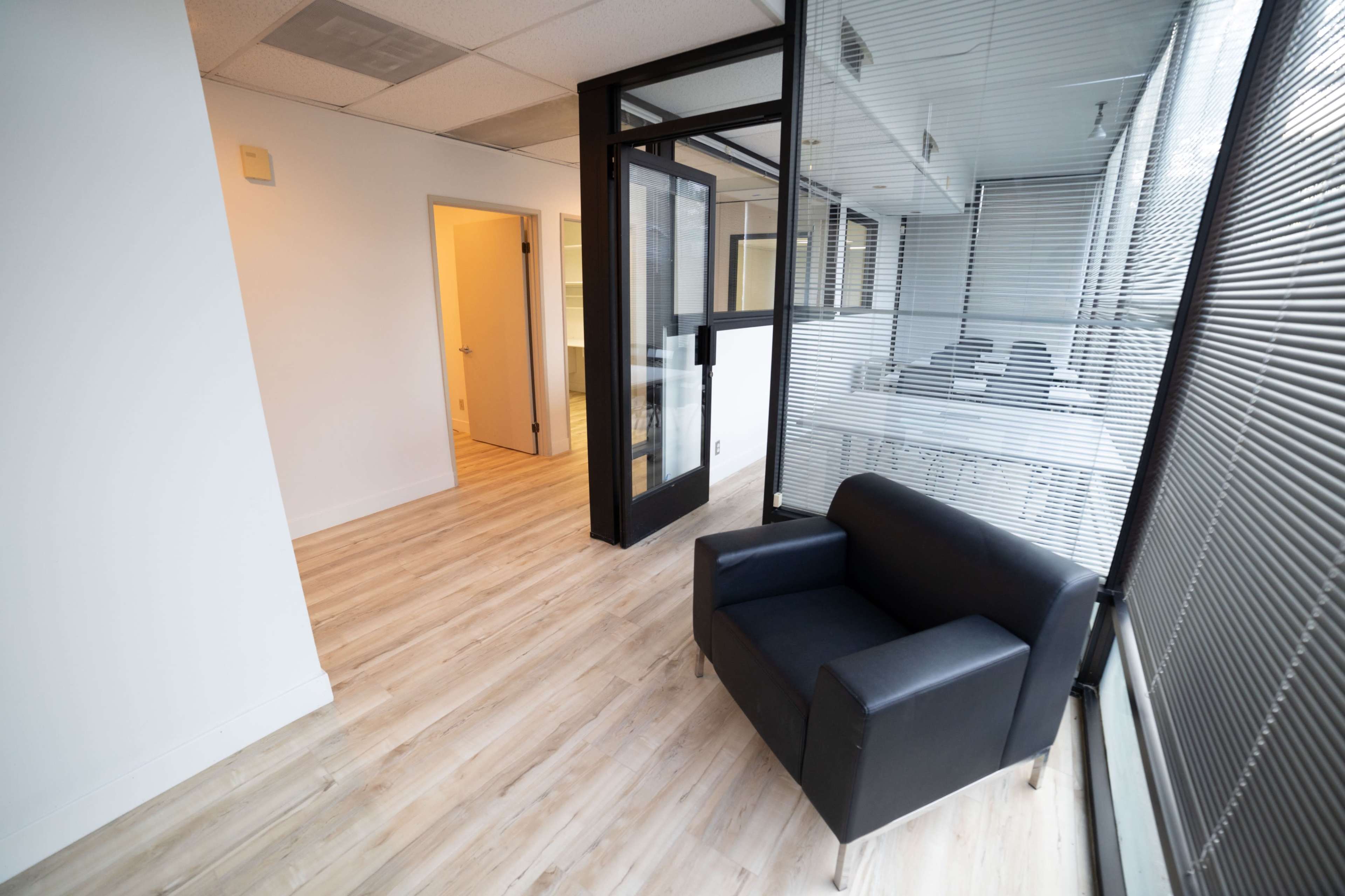 A minimalistic office space featuring a black armchair next to a doorway, with large windows and wooden flooring.