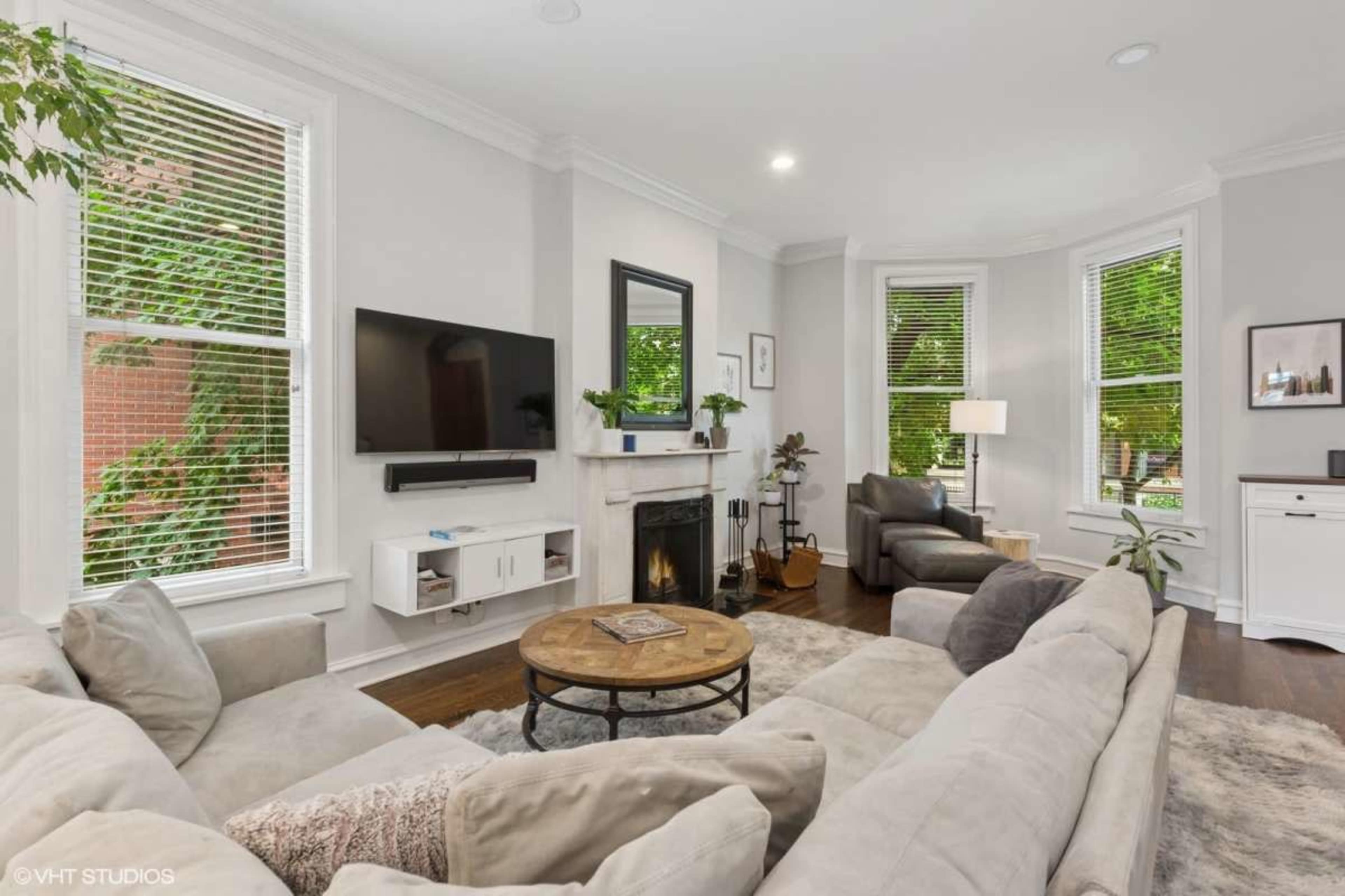 The image shows a cozy living room featuring a sectional sofa, a television mounted on the wall, a fireplace, and large windows with natural light.
