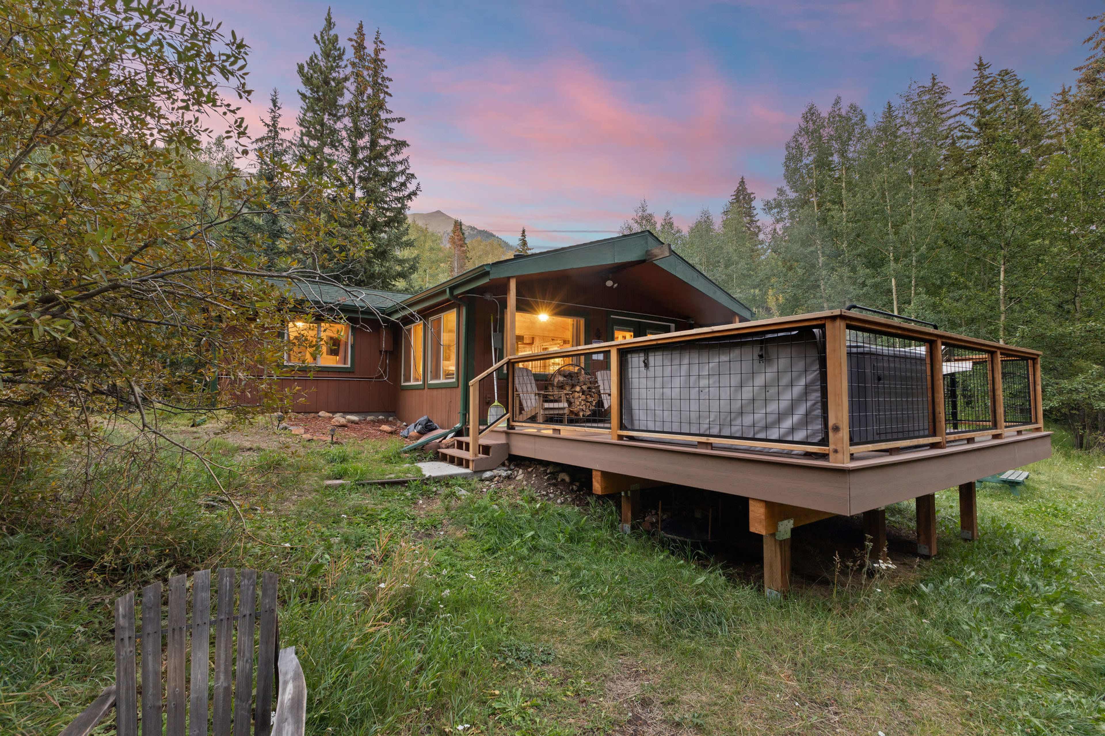 A wooden cabin with a deck is surrounded by trees and grass under a colorful sunset sky.