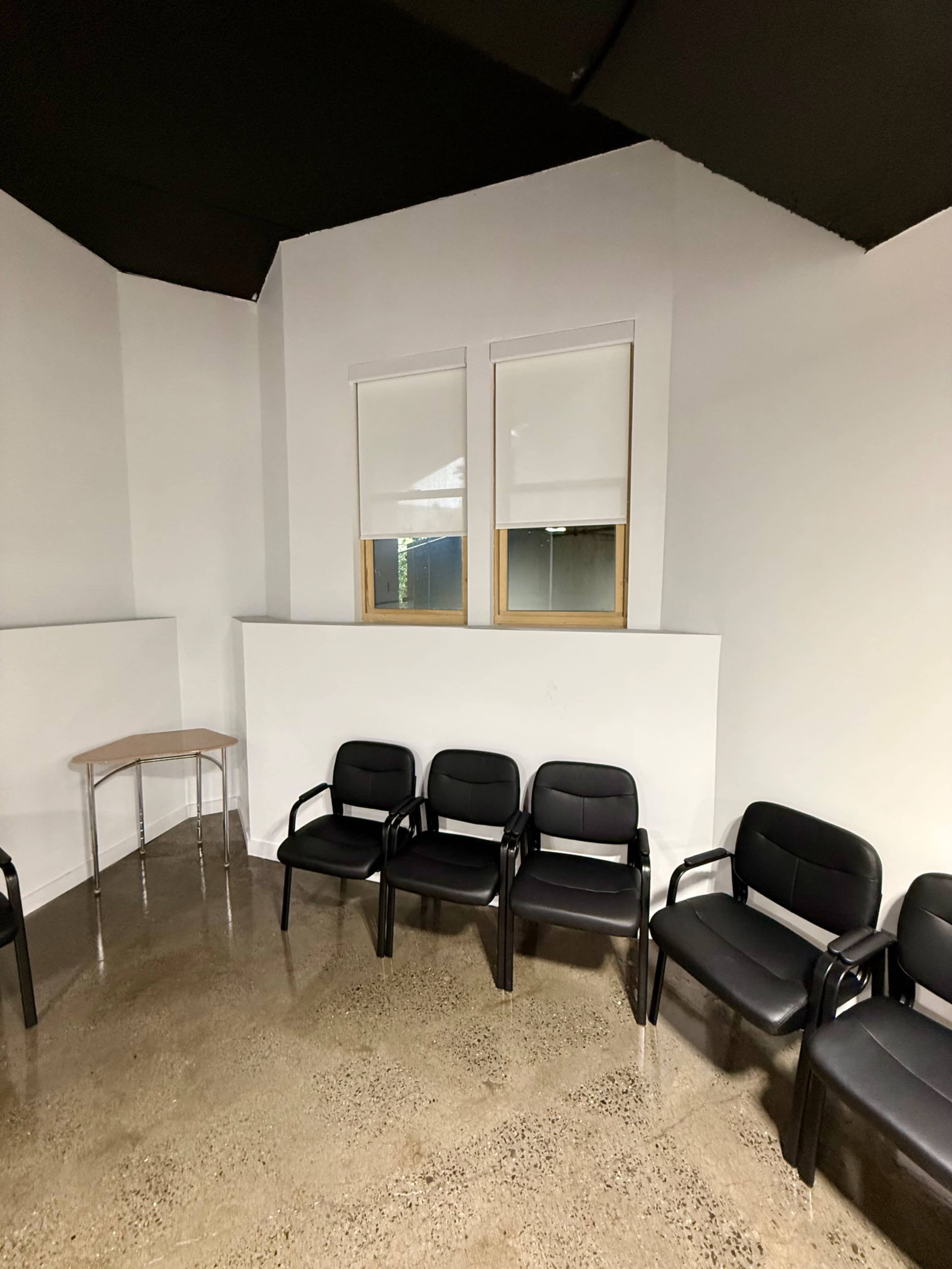 The image shows a simple waiting area with several black chairs arranged along a wall and a small table in one corner.
