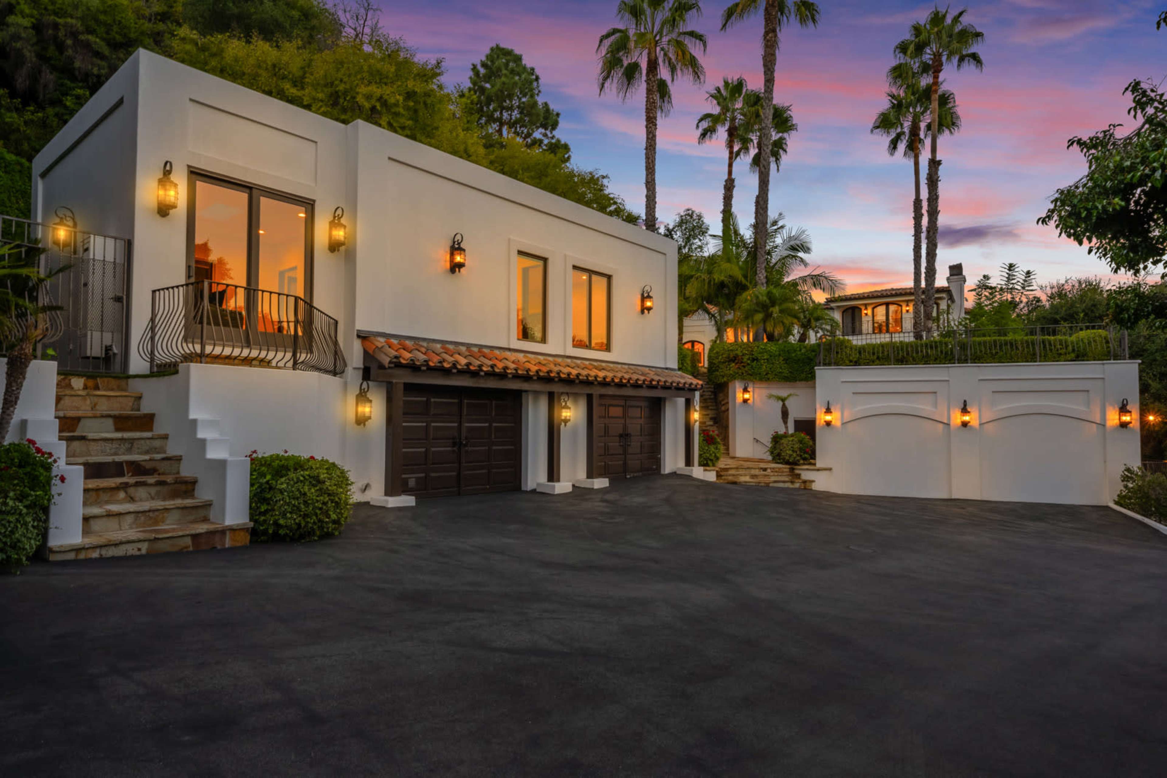 A modern, white house with a terracotta roof, a paved driveway, and palm trees, illuminated by outdoor lighting at dusk.