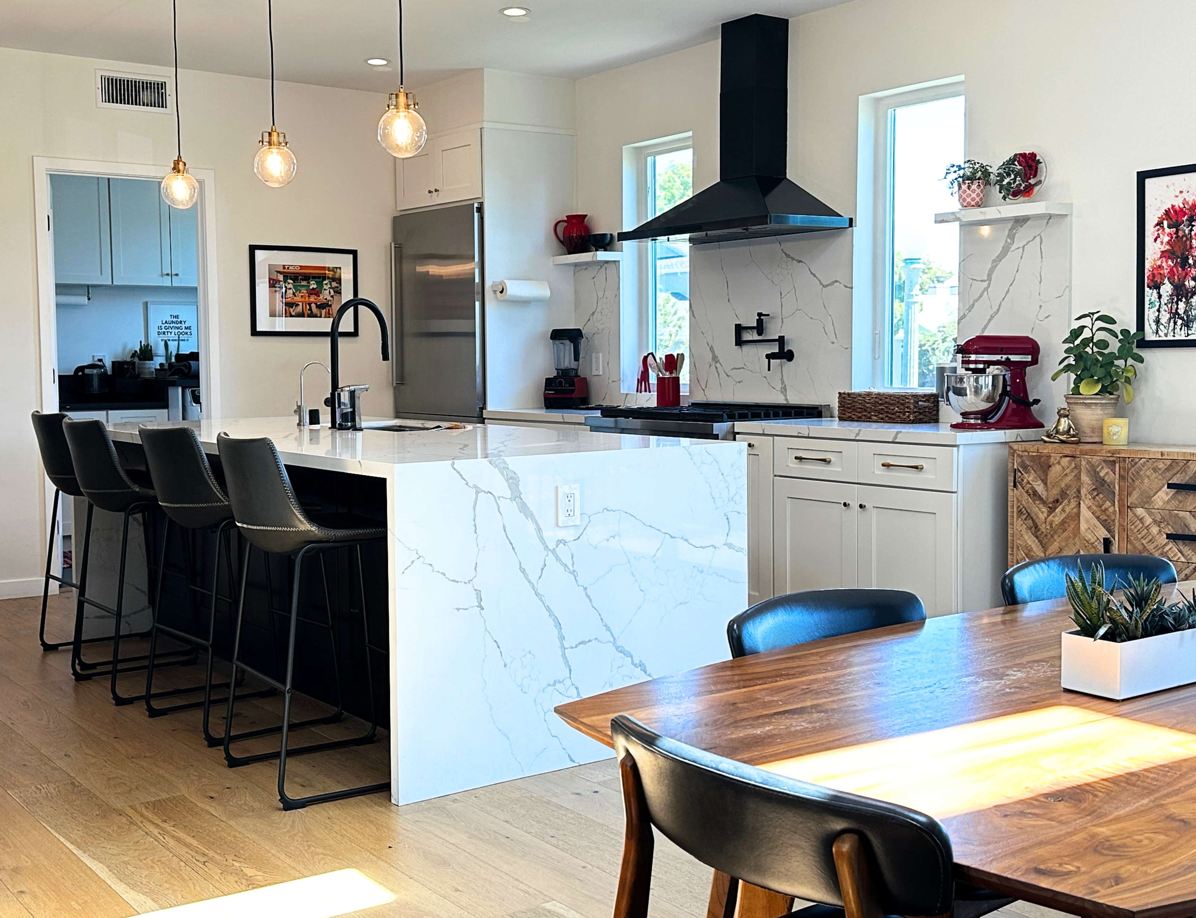 The image shows a modern kitchen area featuring a large island with bar stools, sleek cabinetry, and a dining table beside it.