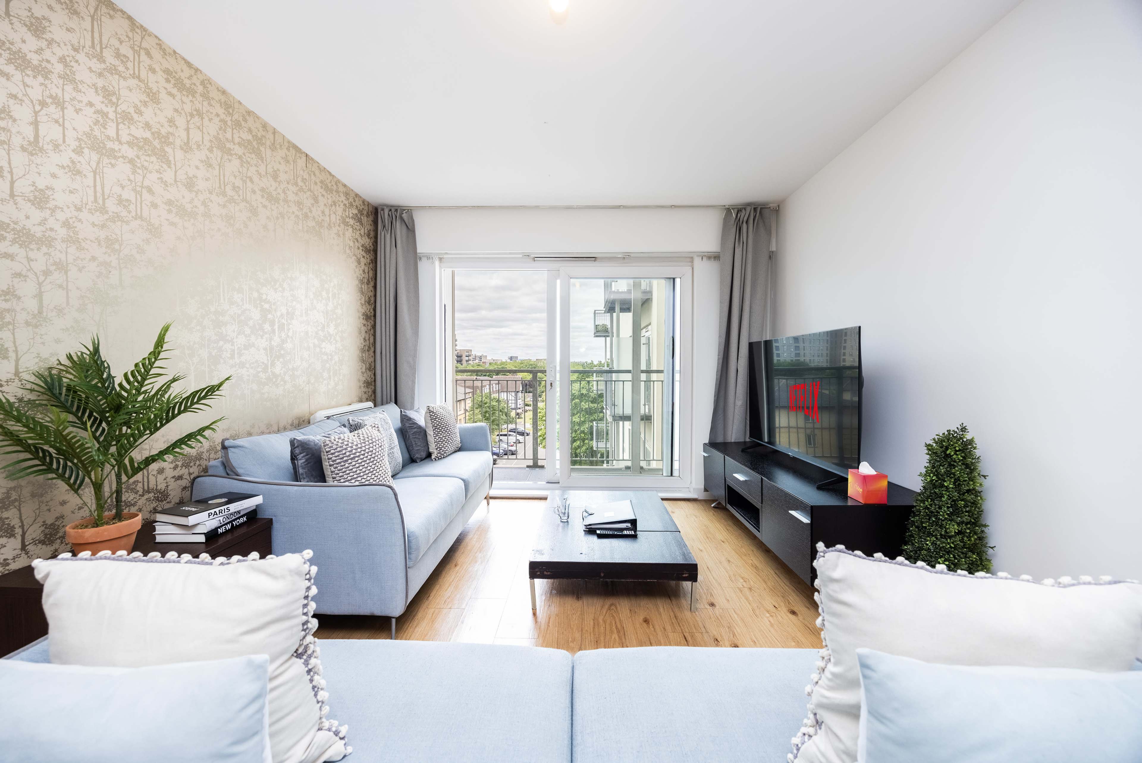 A modern living room features a light blue sofa, a wooden coffee table, and a television against a wall with a large window overlooking a cityscape.