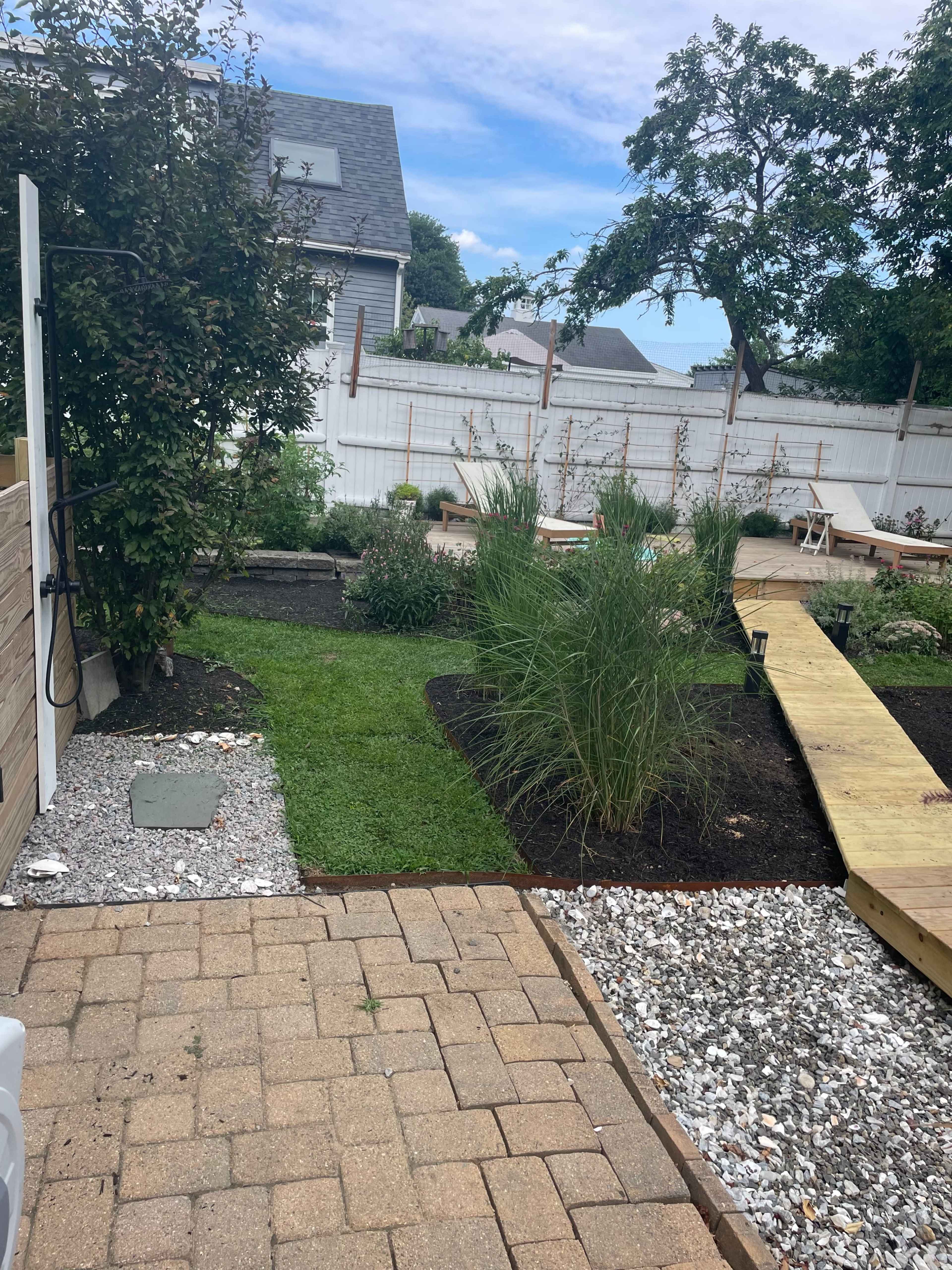 The image shows a landscaped garden featuring a wooden walkway, grassy areas, and various plants, bordered by gravel and a white fence.