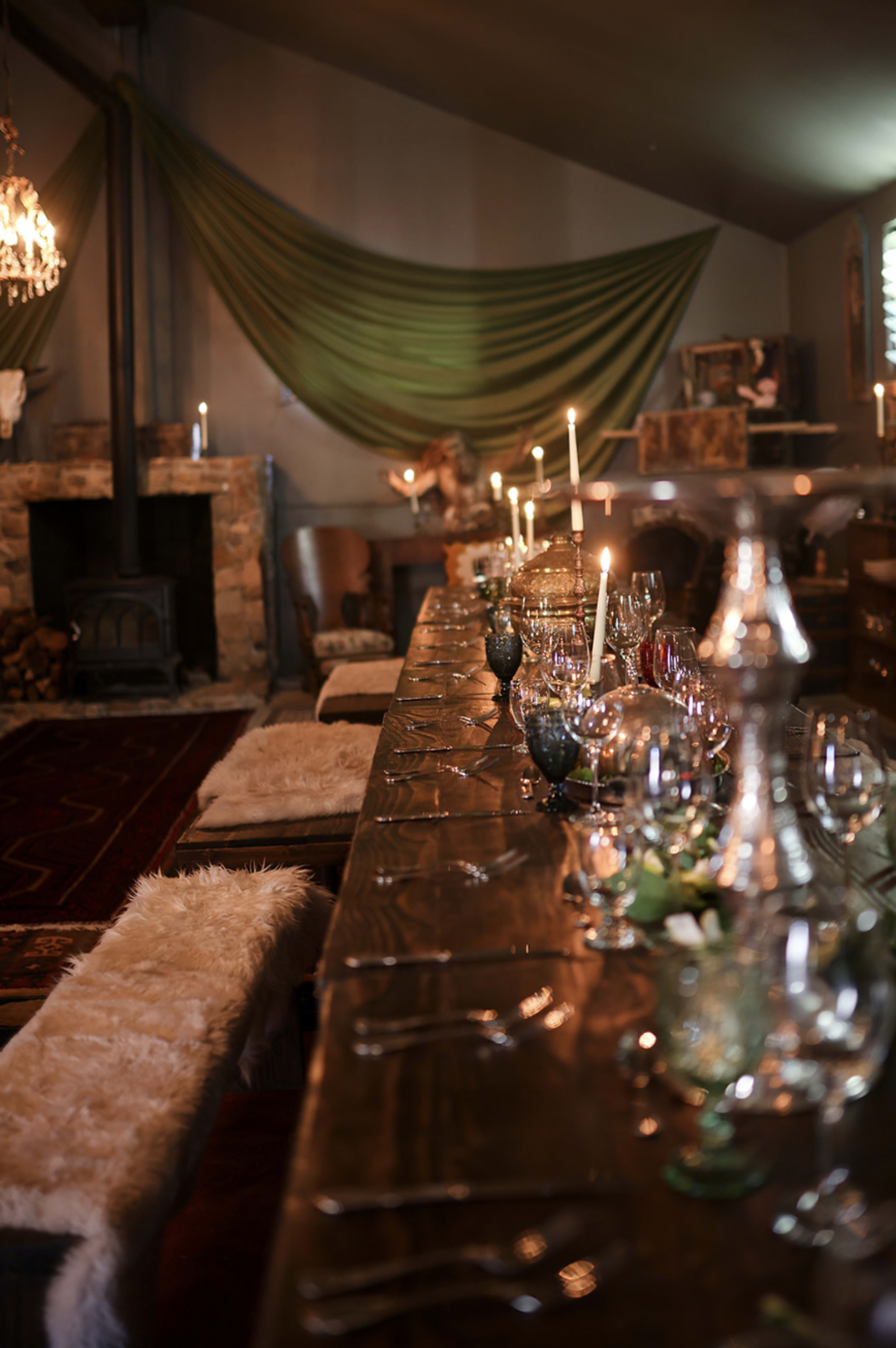 The image shows a long wooden dining table set with glassware and cutlery, surrounded by chairs, with candles lit and a stone fireplace in the background.