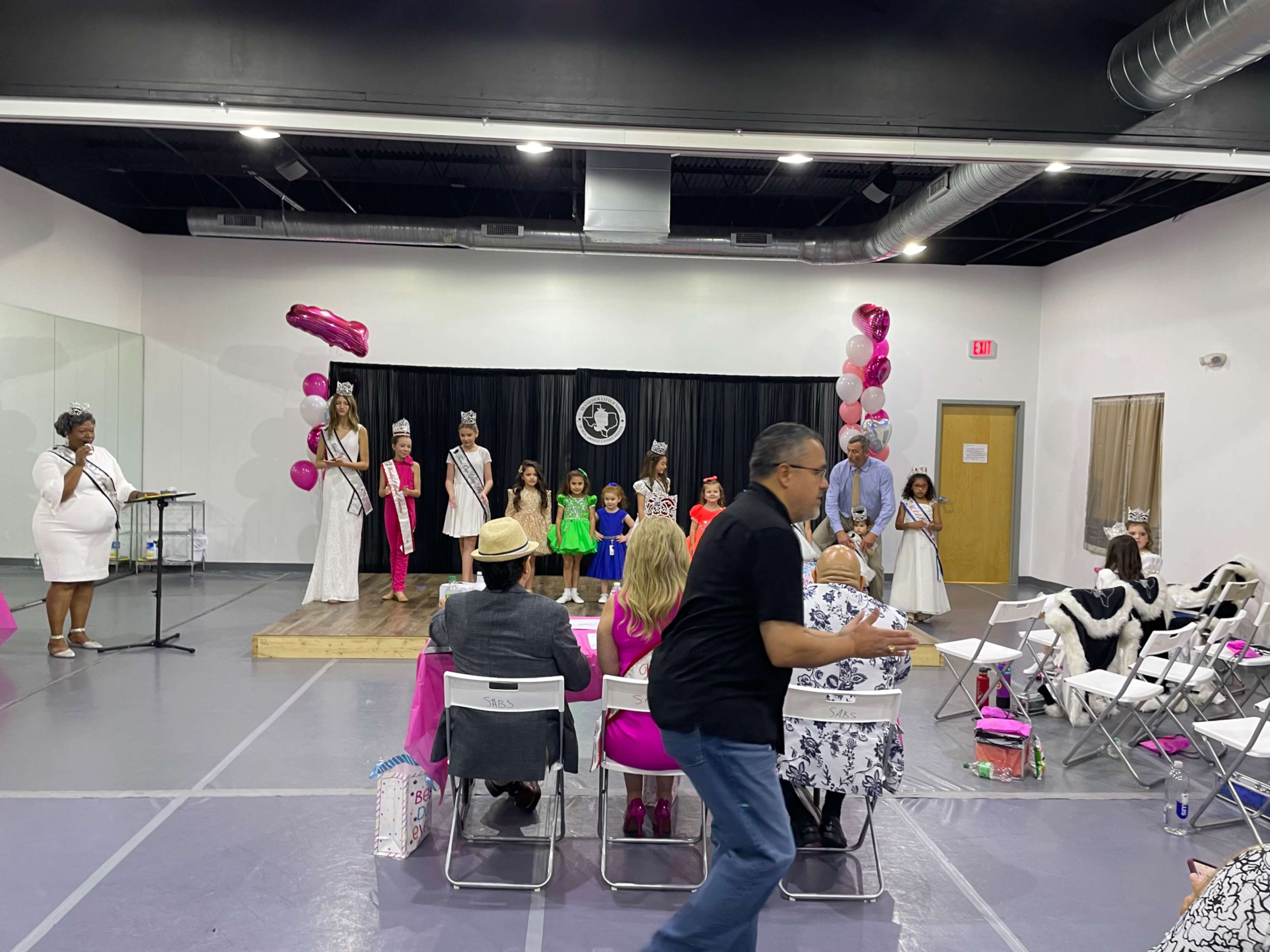 A stage features contestants in formal attire while judges observe from tables, and an audience watches the event in a large room decorated with balloons.