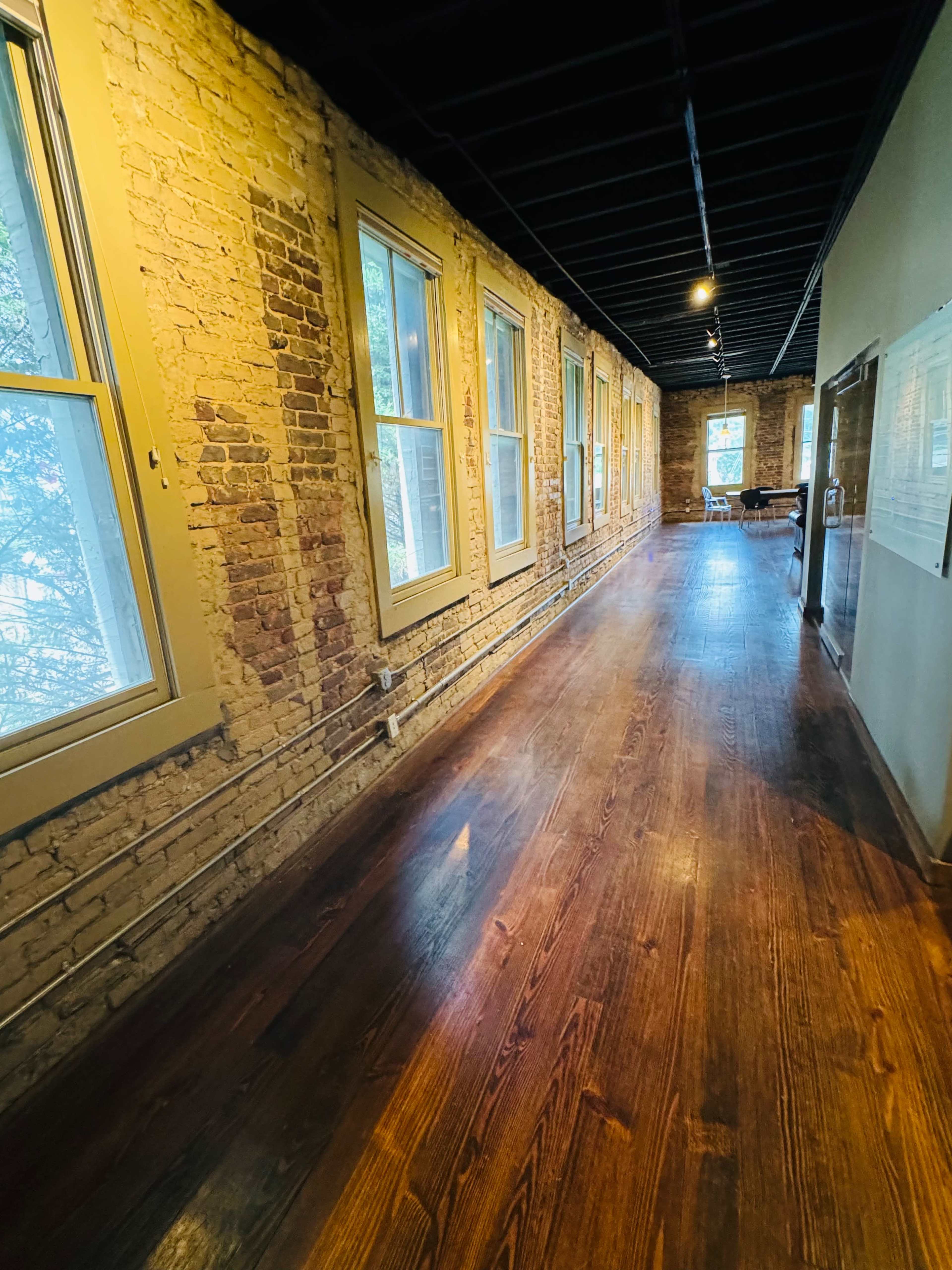 A long, narrow hallway with exposed brick walls, large windows, and wooden flooring.