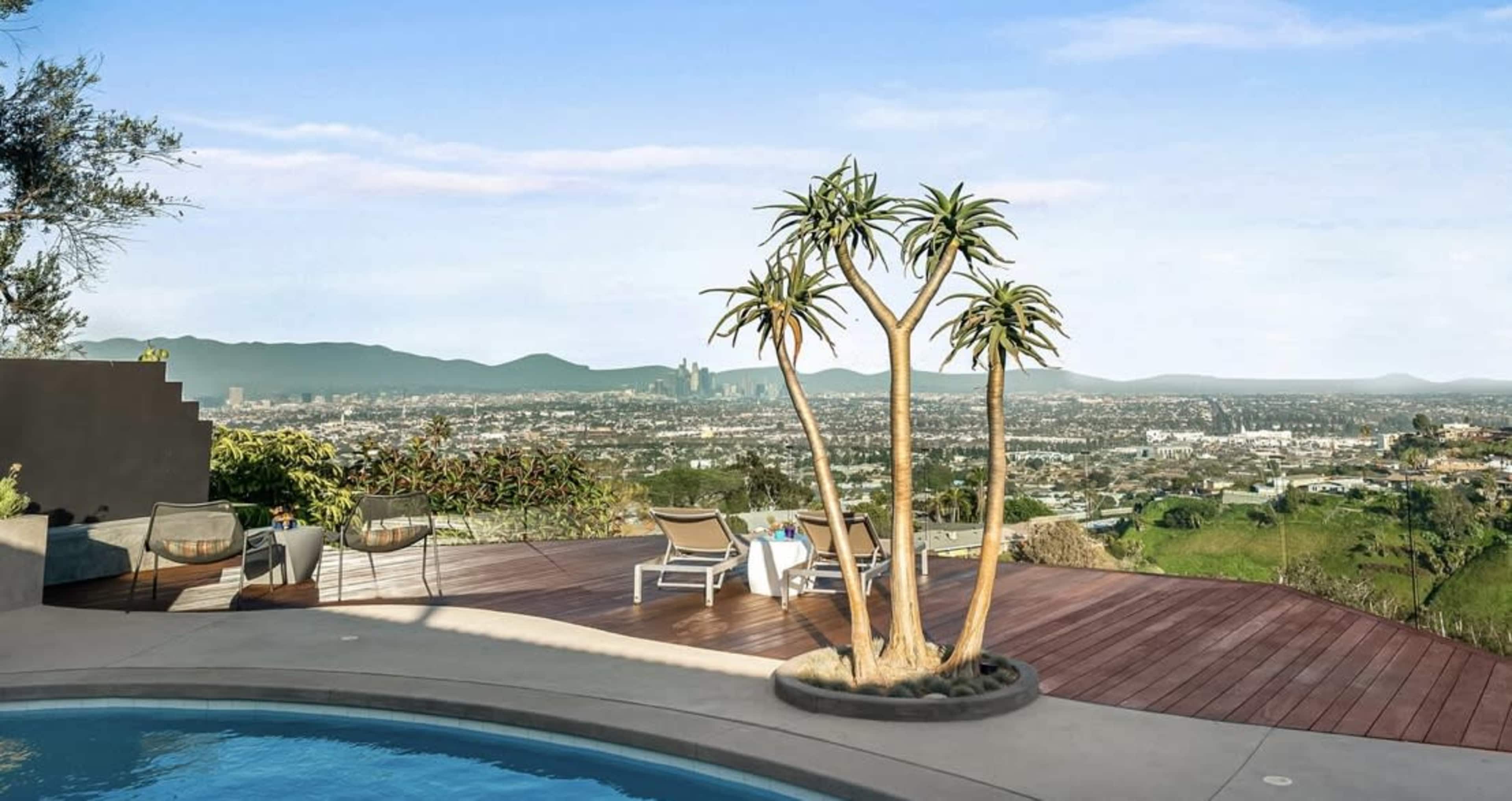A modern outdoor patio features a swimming pool, lounge chairs, and tall potted plants, overlooking a sprawling cityscape and mountains in the distance.