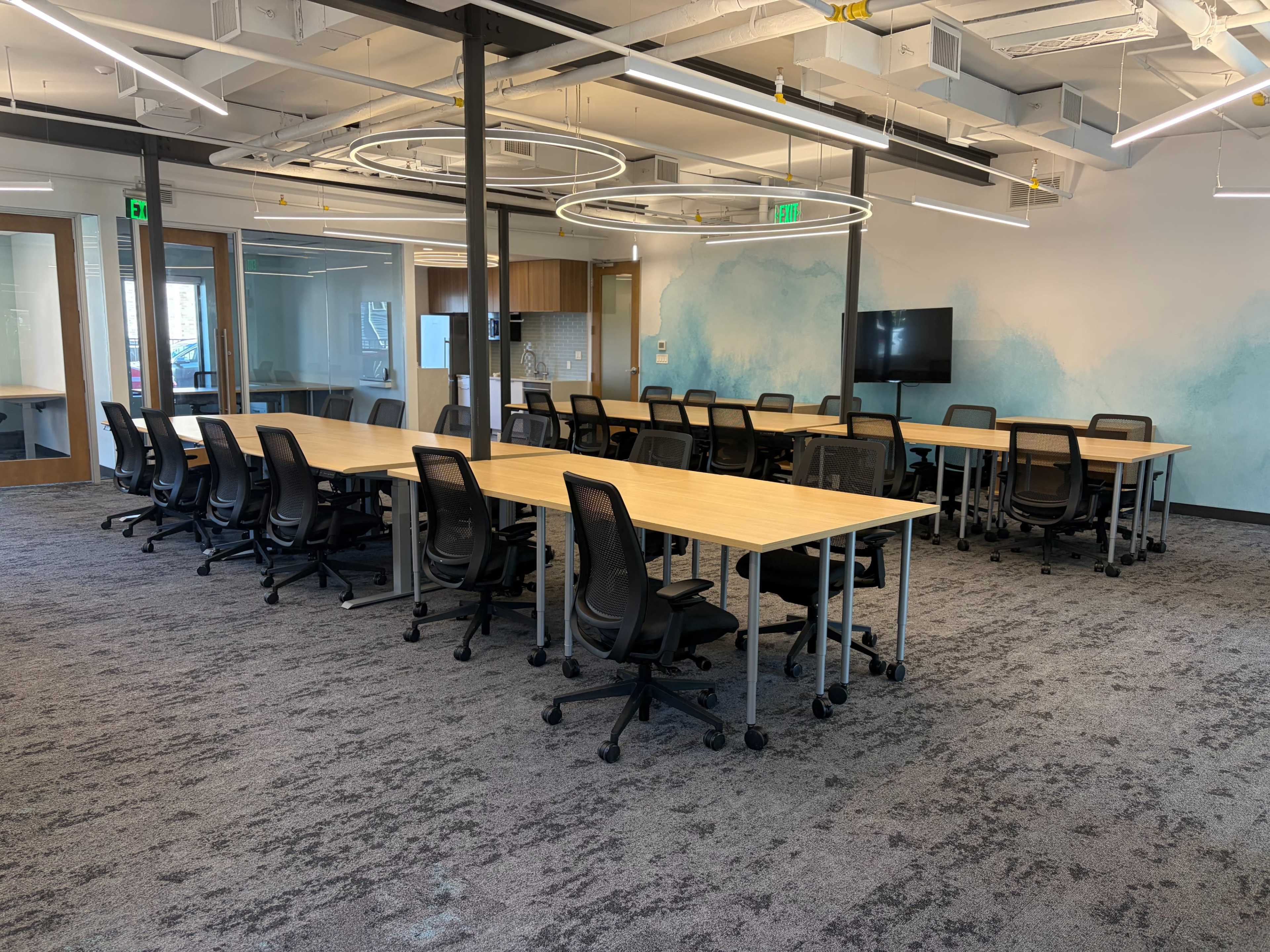 The image shows a modern office space featuring several long tables arranged with black chairs, under circular light fixtures, with walls that display a light blue design.