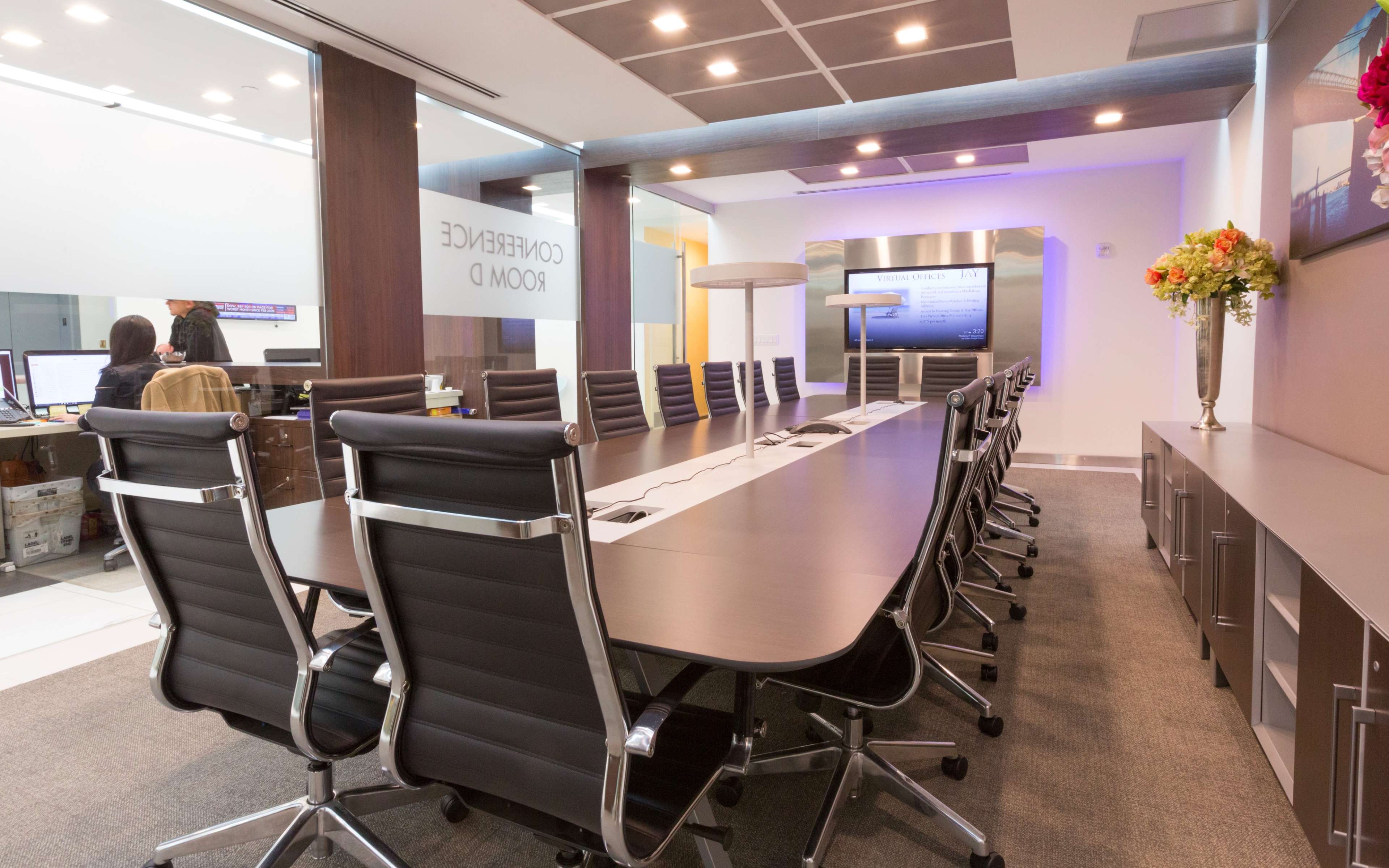 A modern conference room with a long, rectangular table and black ergonomic chairs, complemented by a digital display and decorative flowers.
