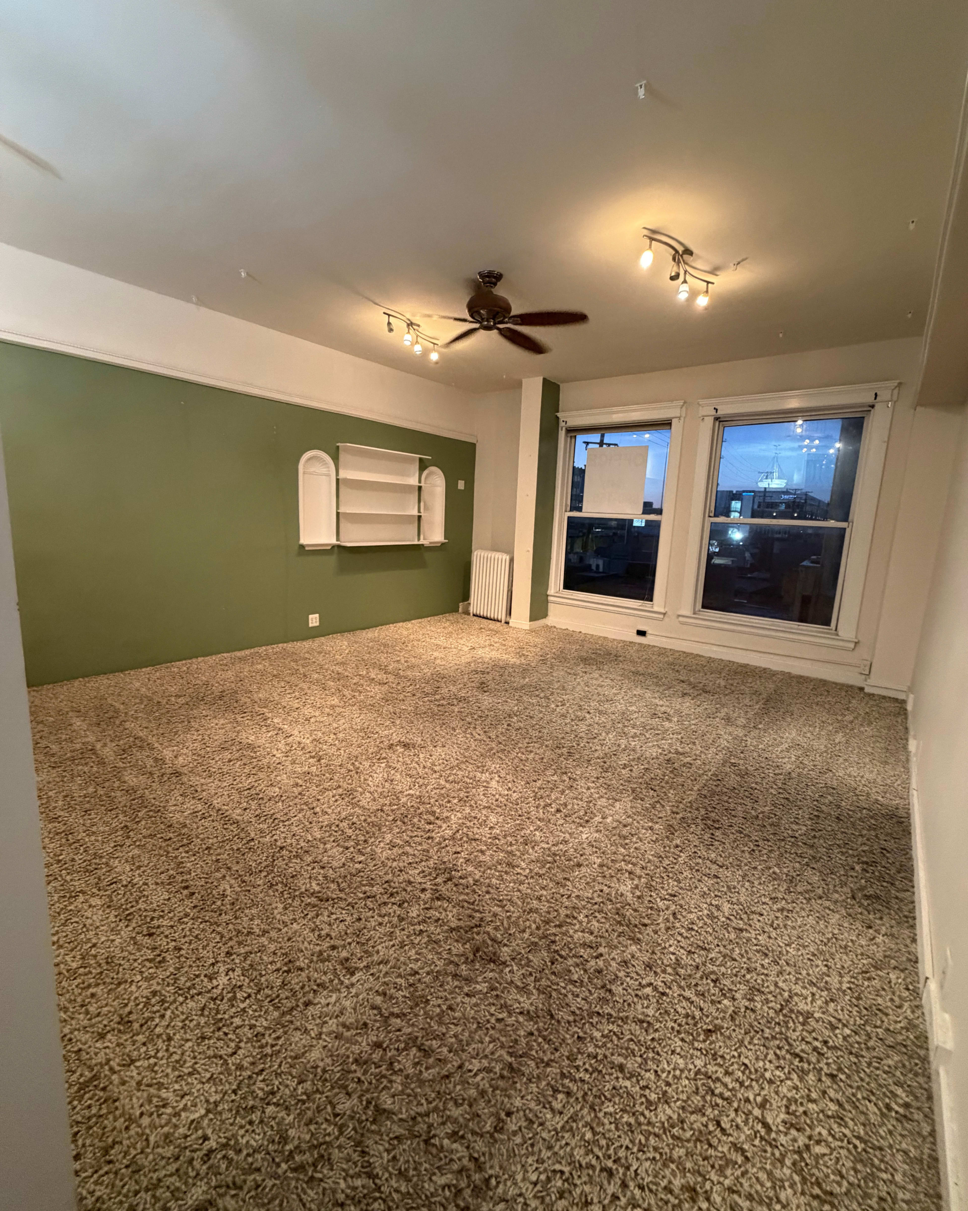 The image shows an empty room with green walls, a ceiling fan, and large windows providing evening light, along with a carpeted floor.