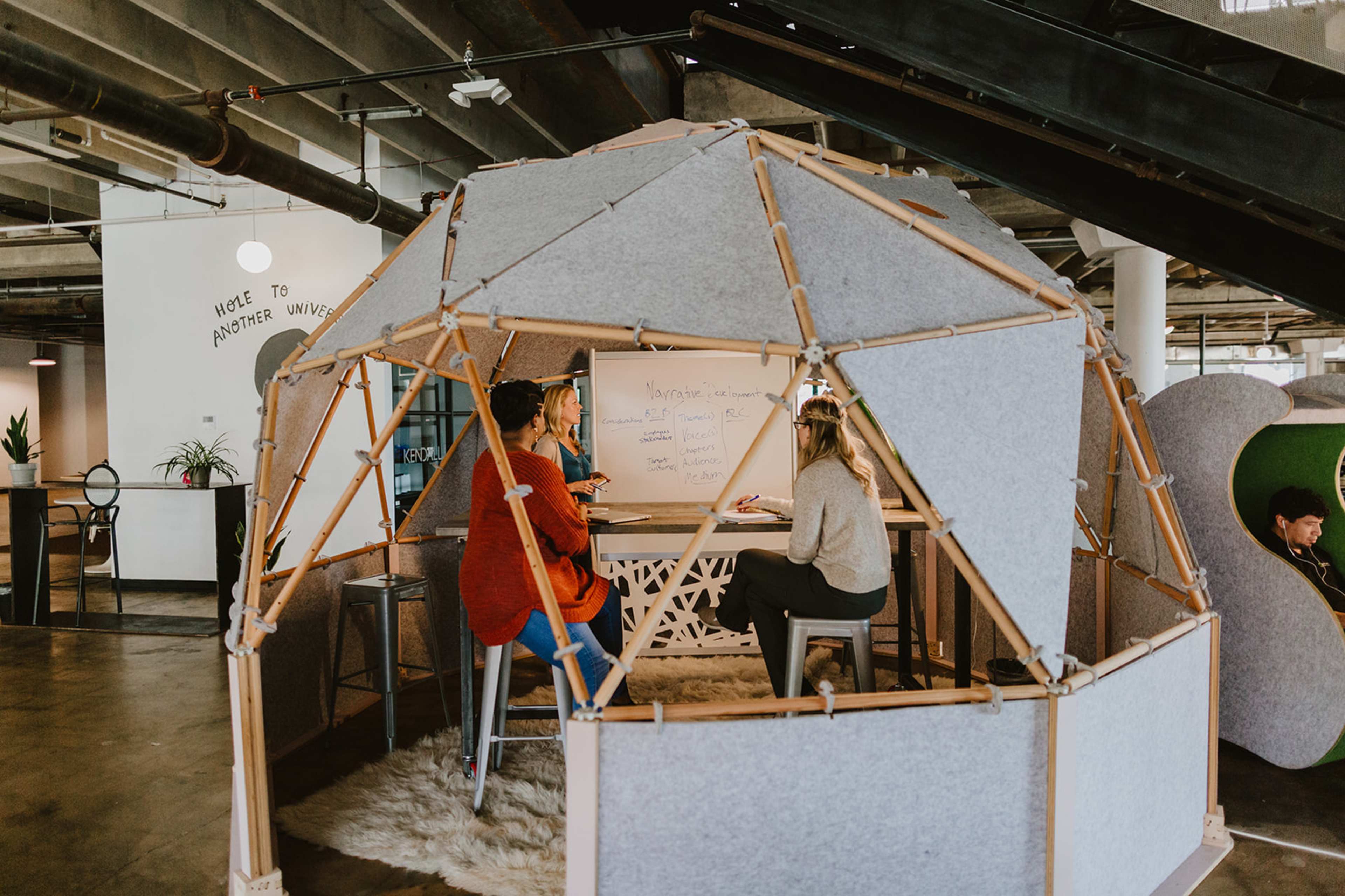 Two people are sitting inside a geodesic dome structure, engaging in a discussion while writing on a whiteboard in a modern office space.