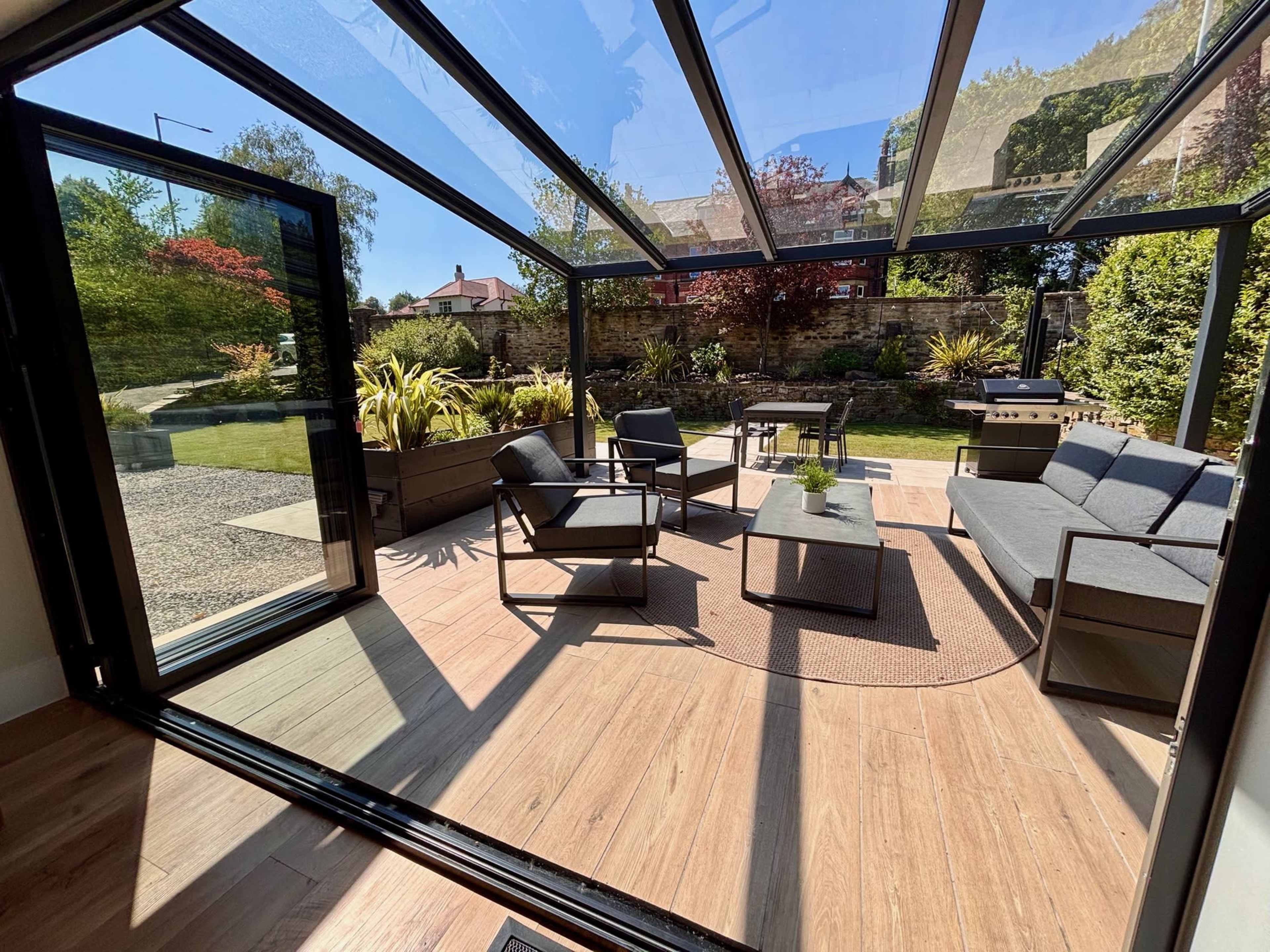 A modern patio area features a glass-roofed structure with seating, a table, and greenery visible through the open doors.