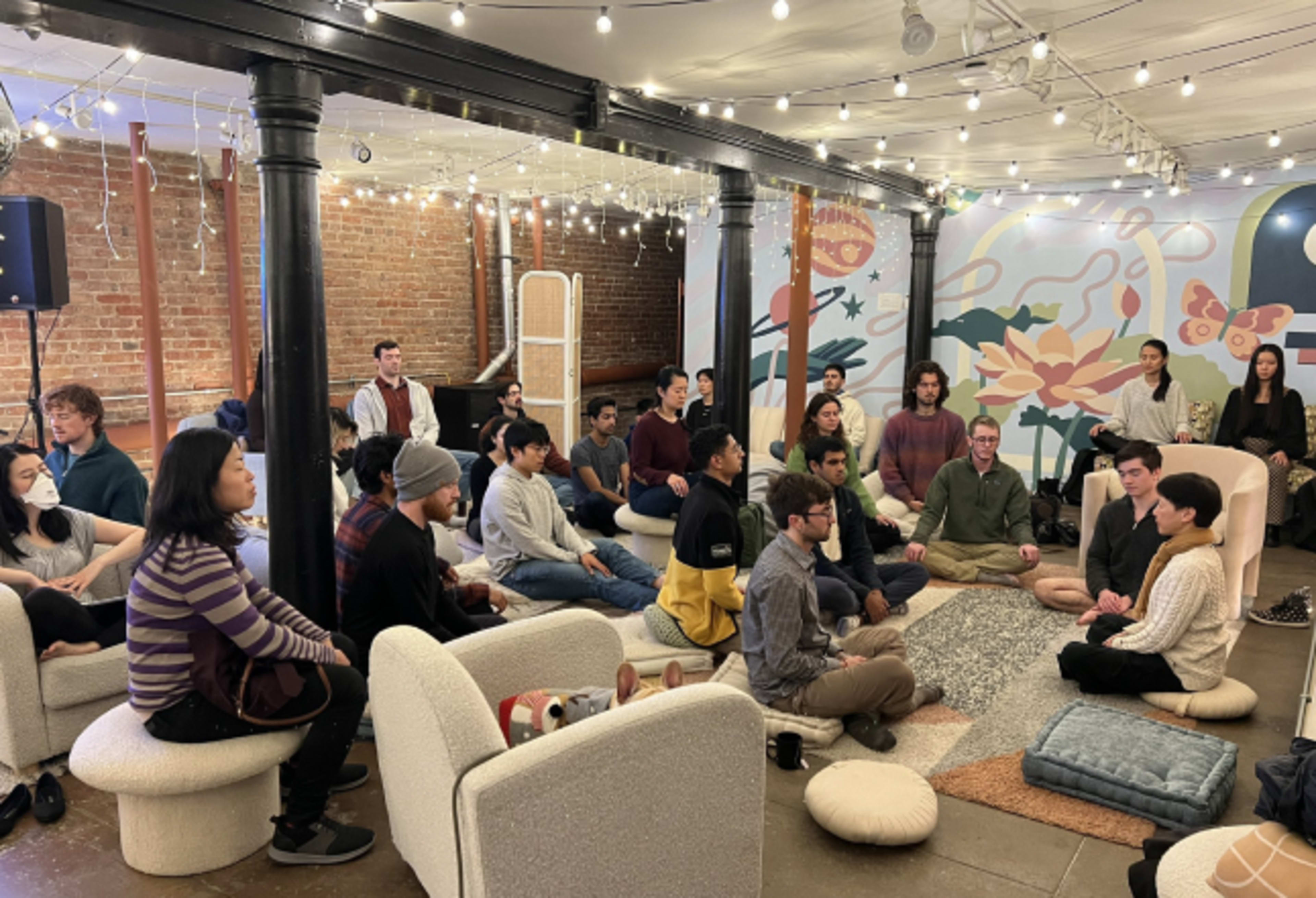 A group of people sits in a circle on elevated platforms in a softly lit room with decorative walls.