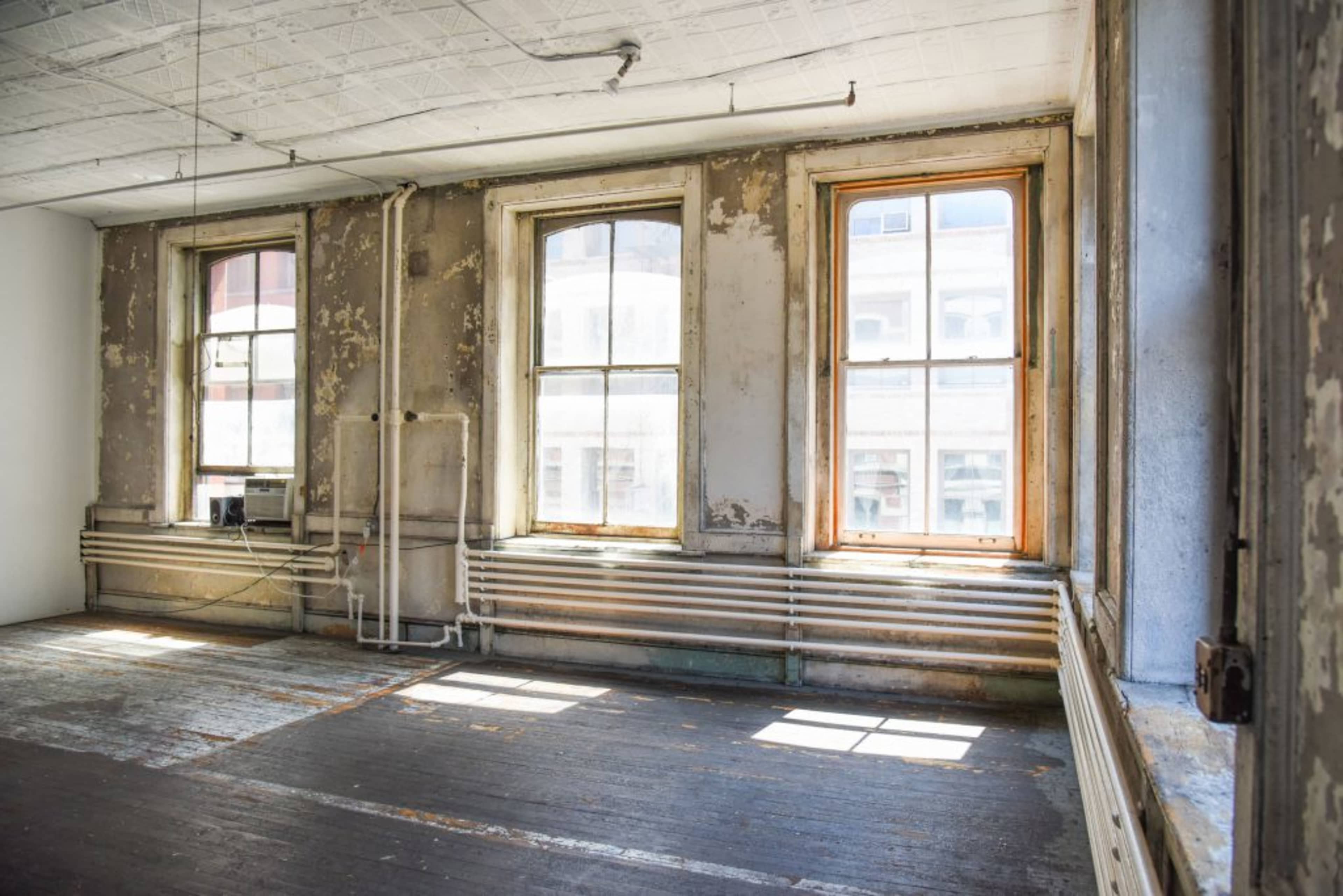 The image shows an empty room with peeling walls, wooden floors, and three large windows overlooking a cityscape.