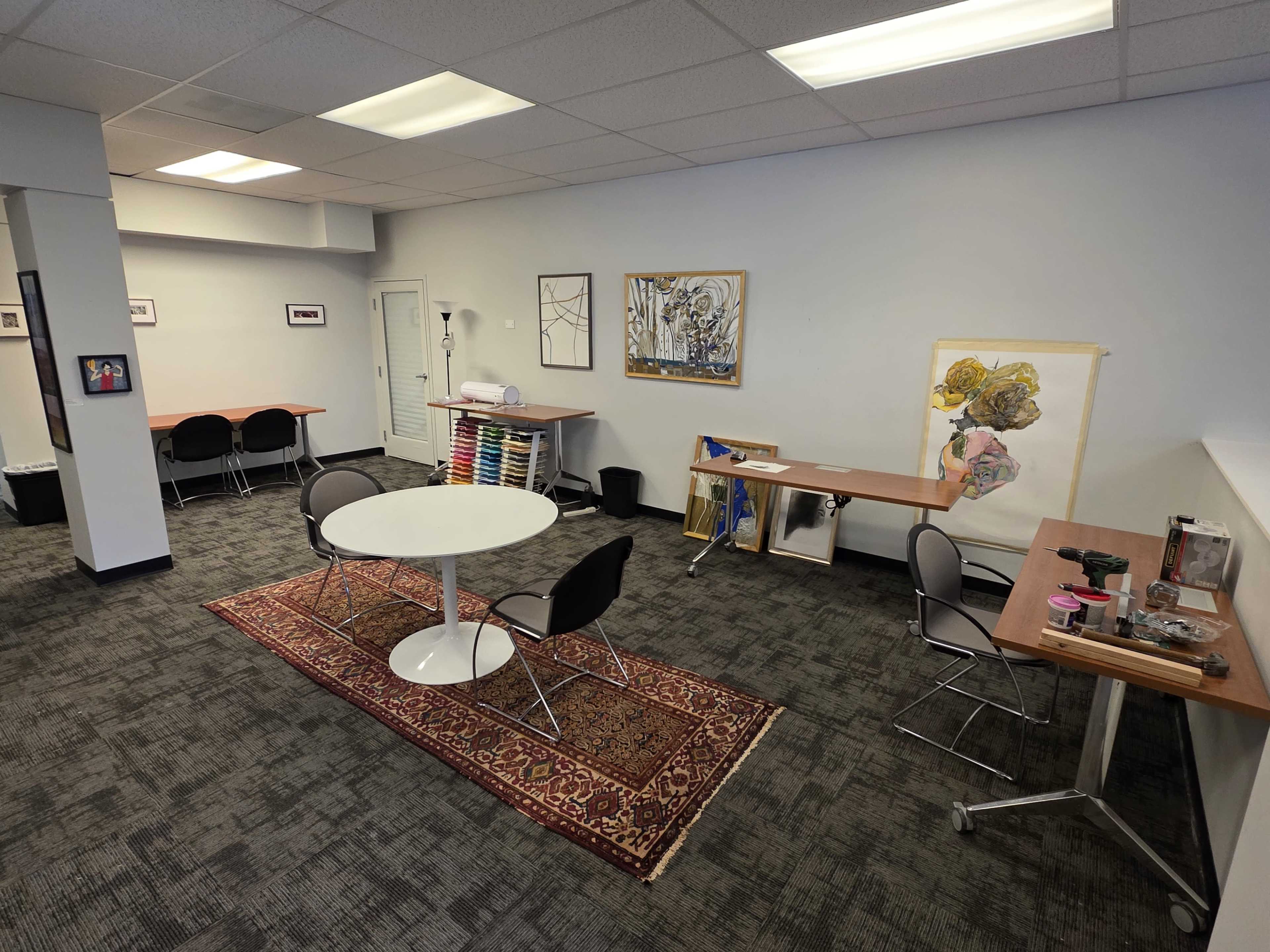 The image shows a spacious, minimalist office with a round table and chairs, artwork on the walls, and a workspace with art supplies.