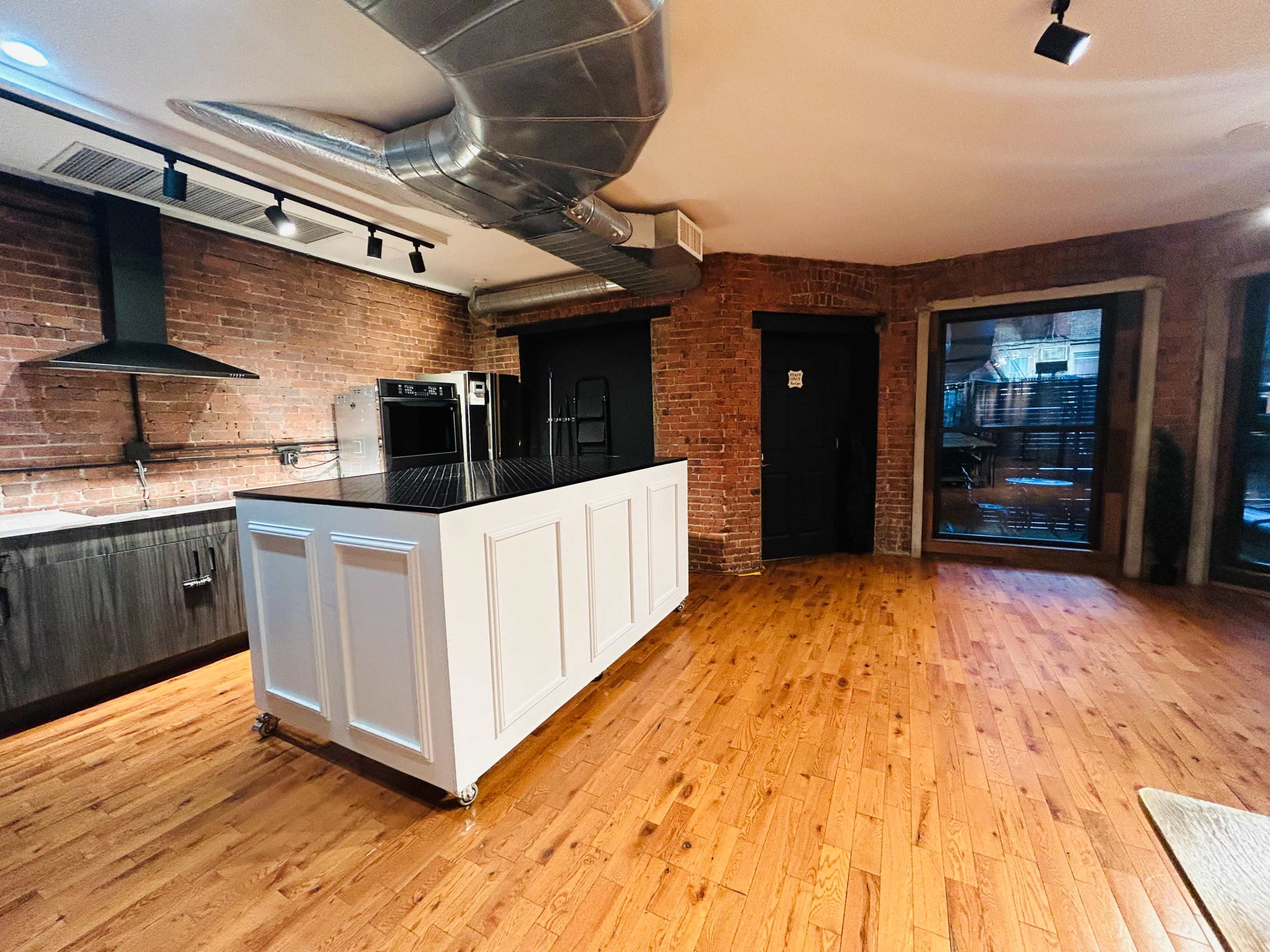 The image shows a modern kitchen with a large island in the center, brick walls, and stainless steel fixtures.