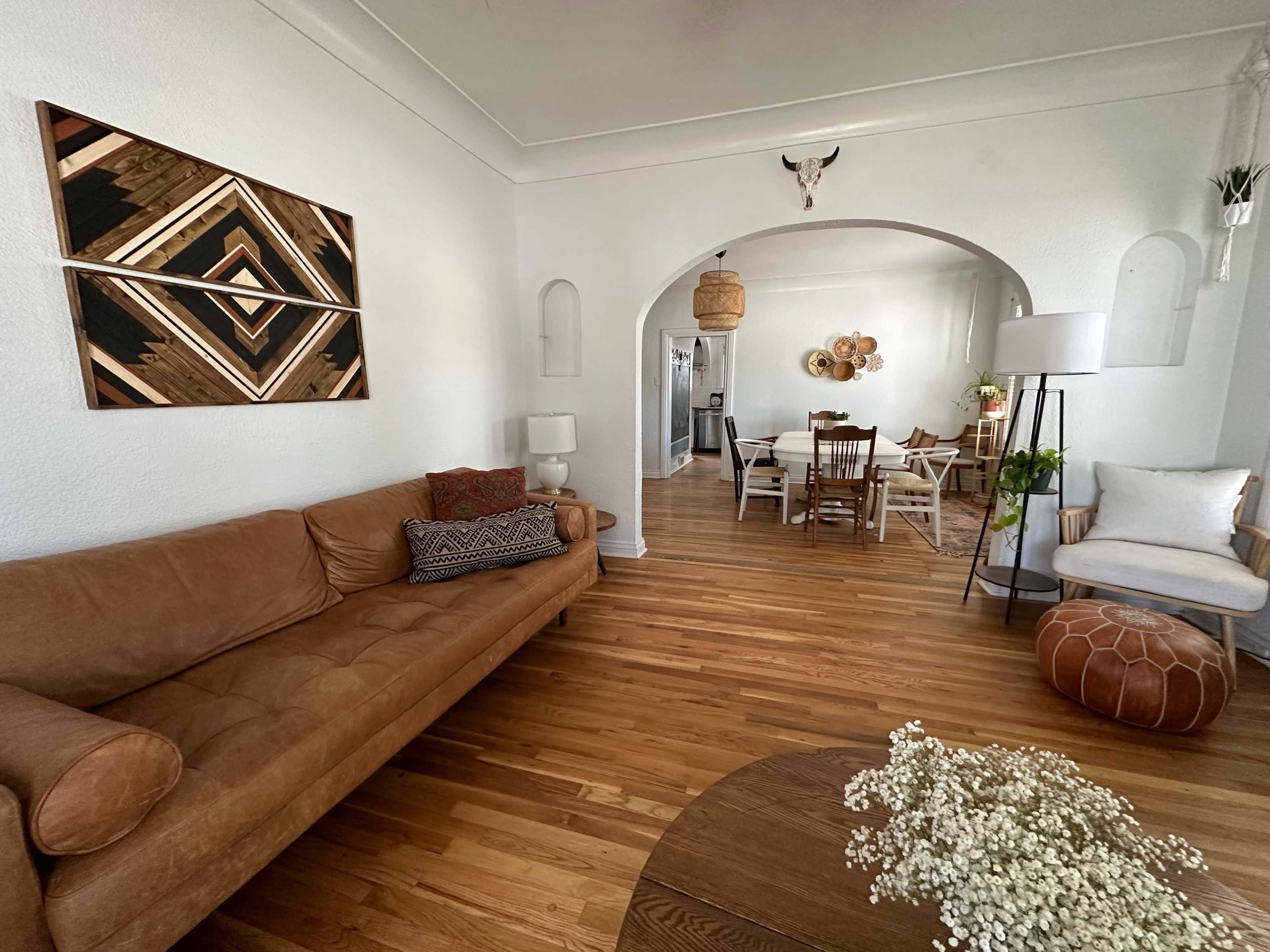 The image shows a living space with a brown leather couch, a wooden coffee table, and a dining area visible through an arched doorway, featuring a modern dining table and chairs.