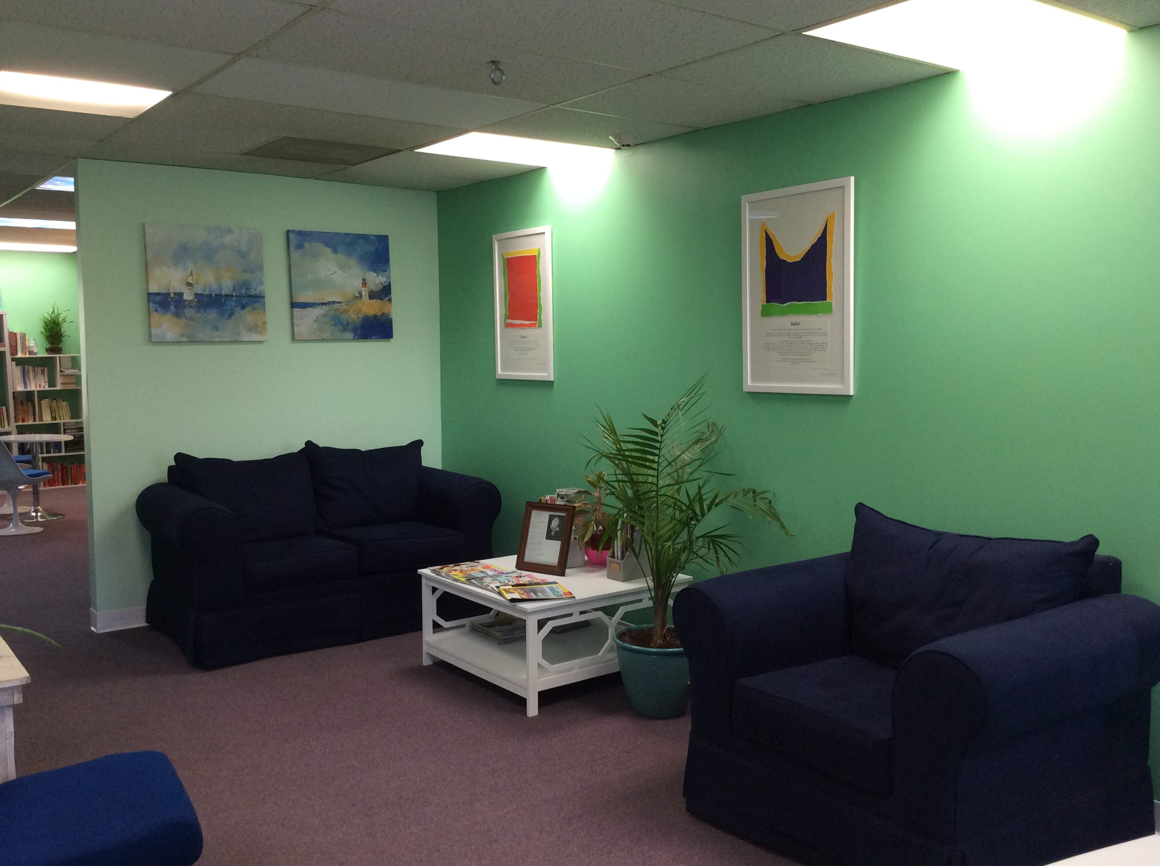 A waiting area with two blue sofas against a green wall, framed artwork, a coffee table with magazines, and a potted plant.