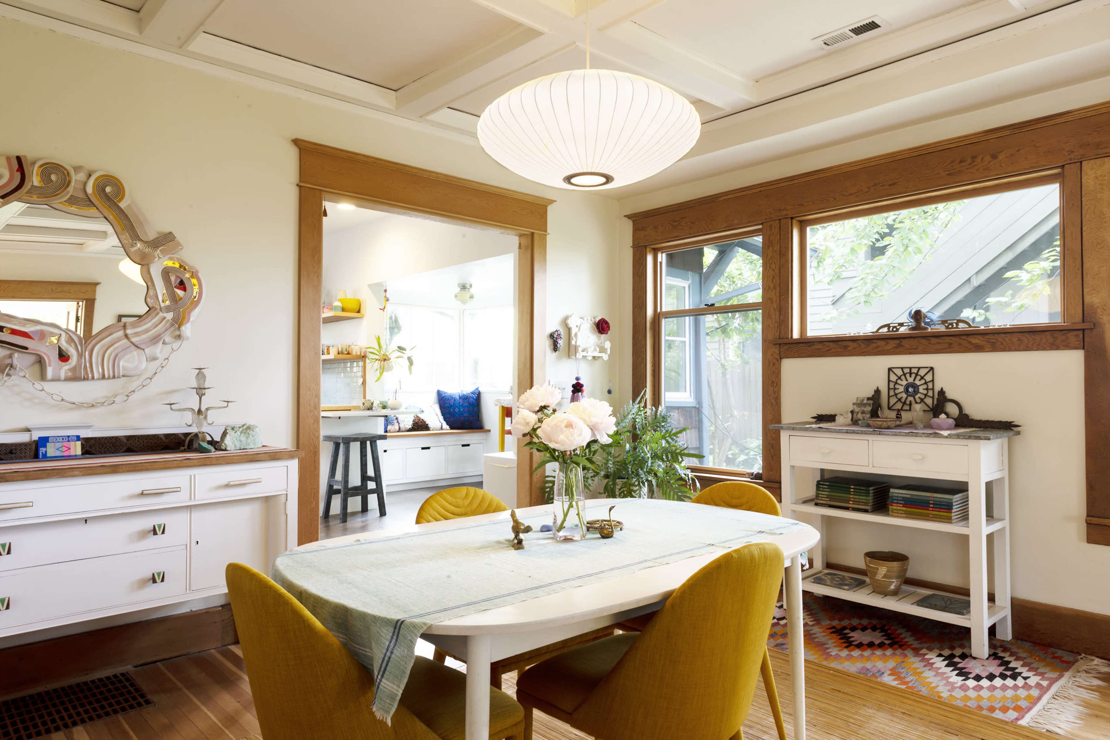 A dining area features a round white table surrounded by yellow chairs, with large windows allowing natural light to illuminate the space.