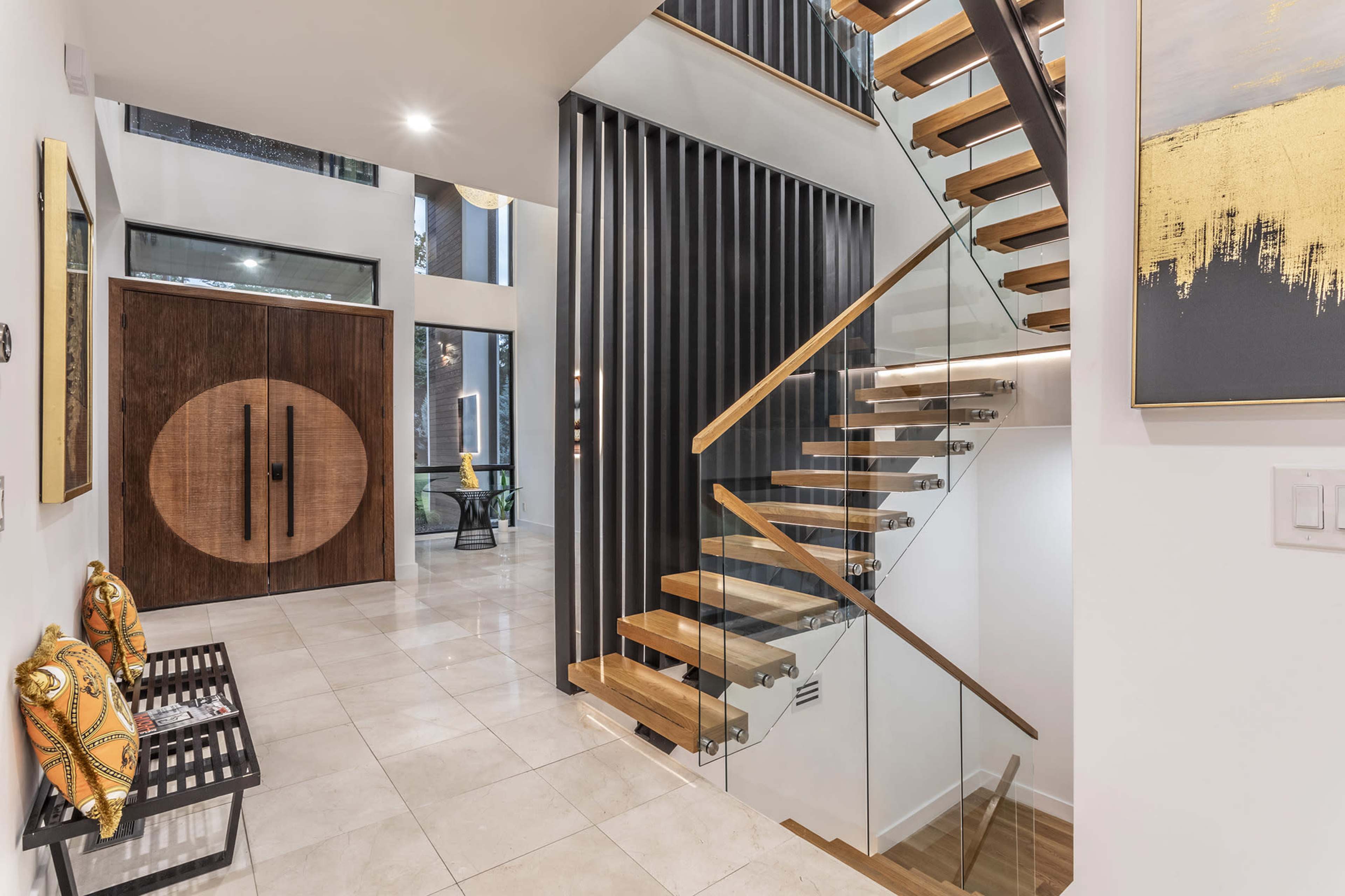 The image shows a modern entryway featuring a wooden staircase with glass railings, tiled floors, and a large circular wooden door.